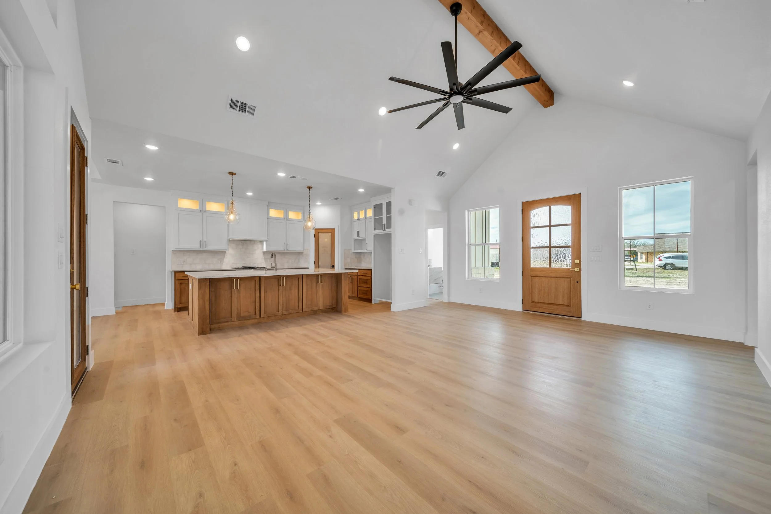 Empty living room with hardwood floors, white walls, large windows, a wooden front door, and an open kitchen area with a central island and pendant lighting.