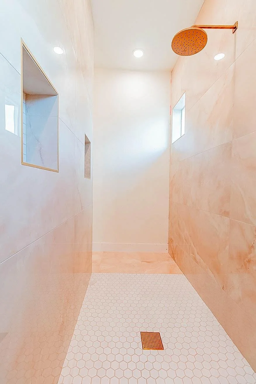A modern shower with peach-colored marble walls, a round overhead shower head, two small square windows, and a white hexagonal tile floor with a gold drain.