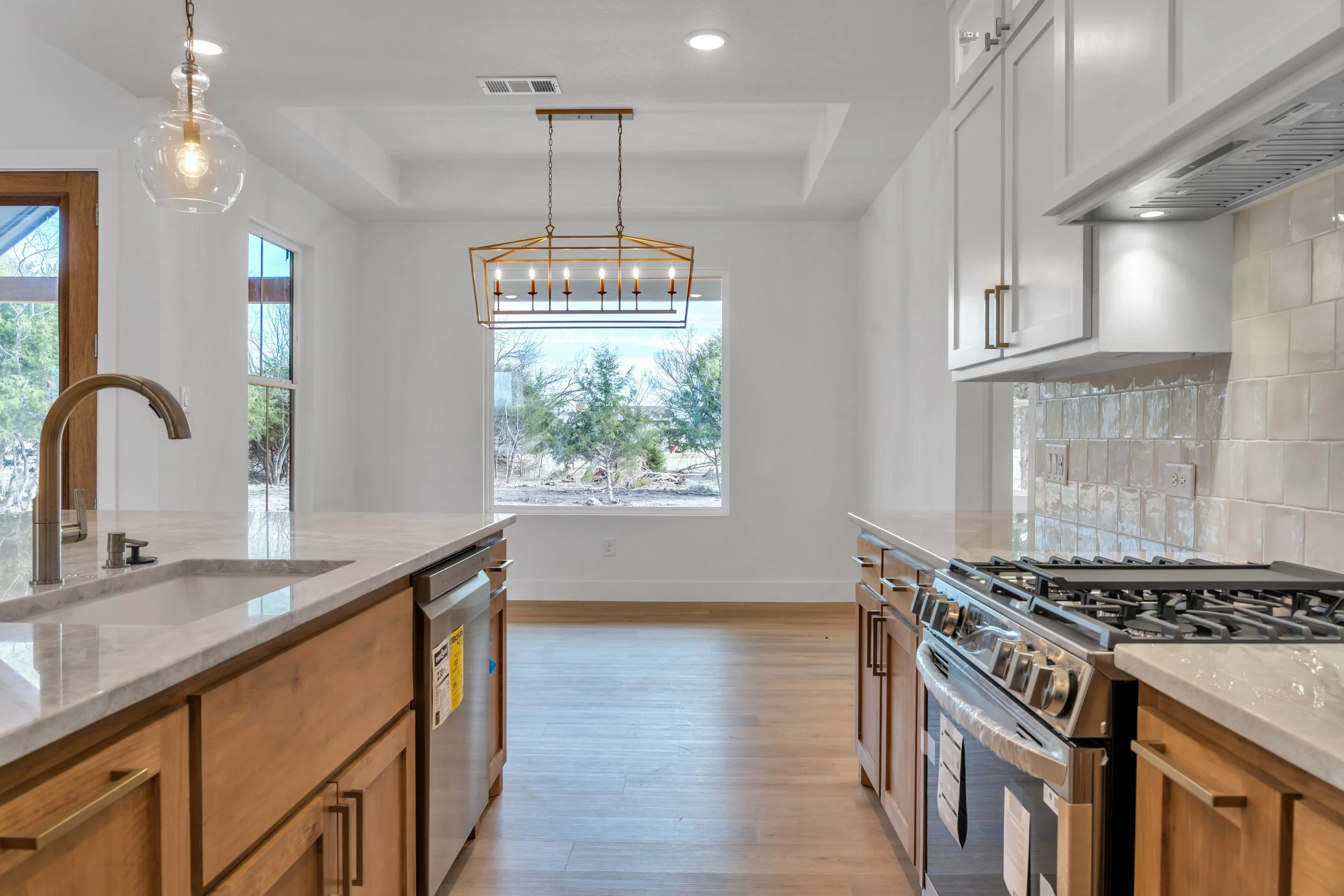 Empty modern kitchen with white walls, wooden cabinets, marble countertops, stainless steel appliances, and a large window showing an outdoor scene with trees.