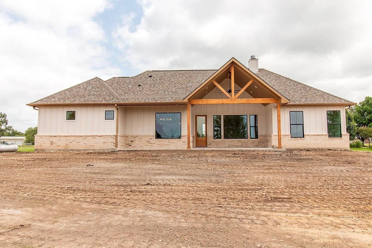 Newly built house in a rural area with a partially finished yard, featuring large windows, a front door, and exposed wooden beams under the gable roof.