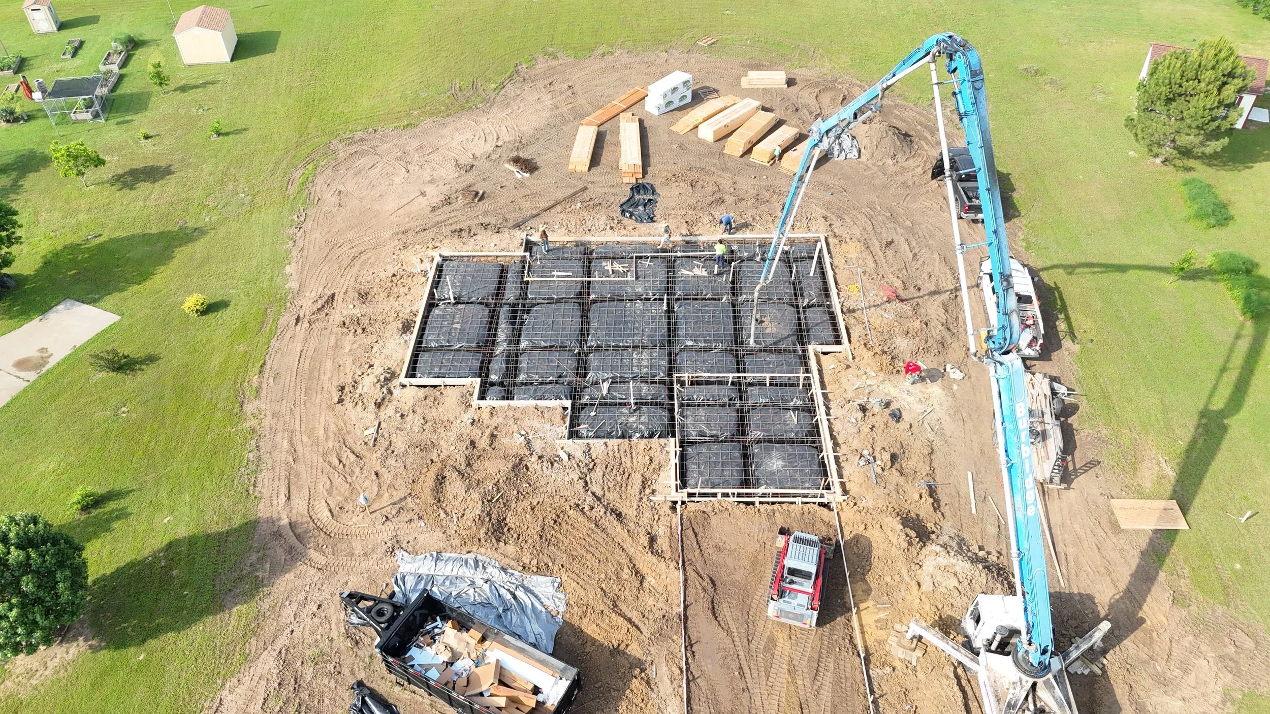 Aerial view of a construction site showing foundation work, concrete pump truck, and workers installing rebar for a building.