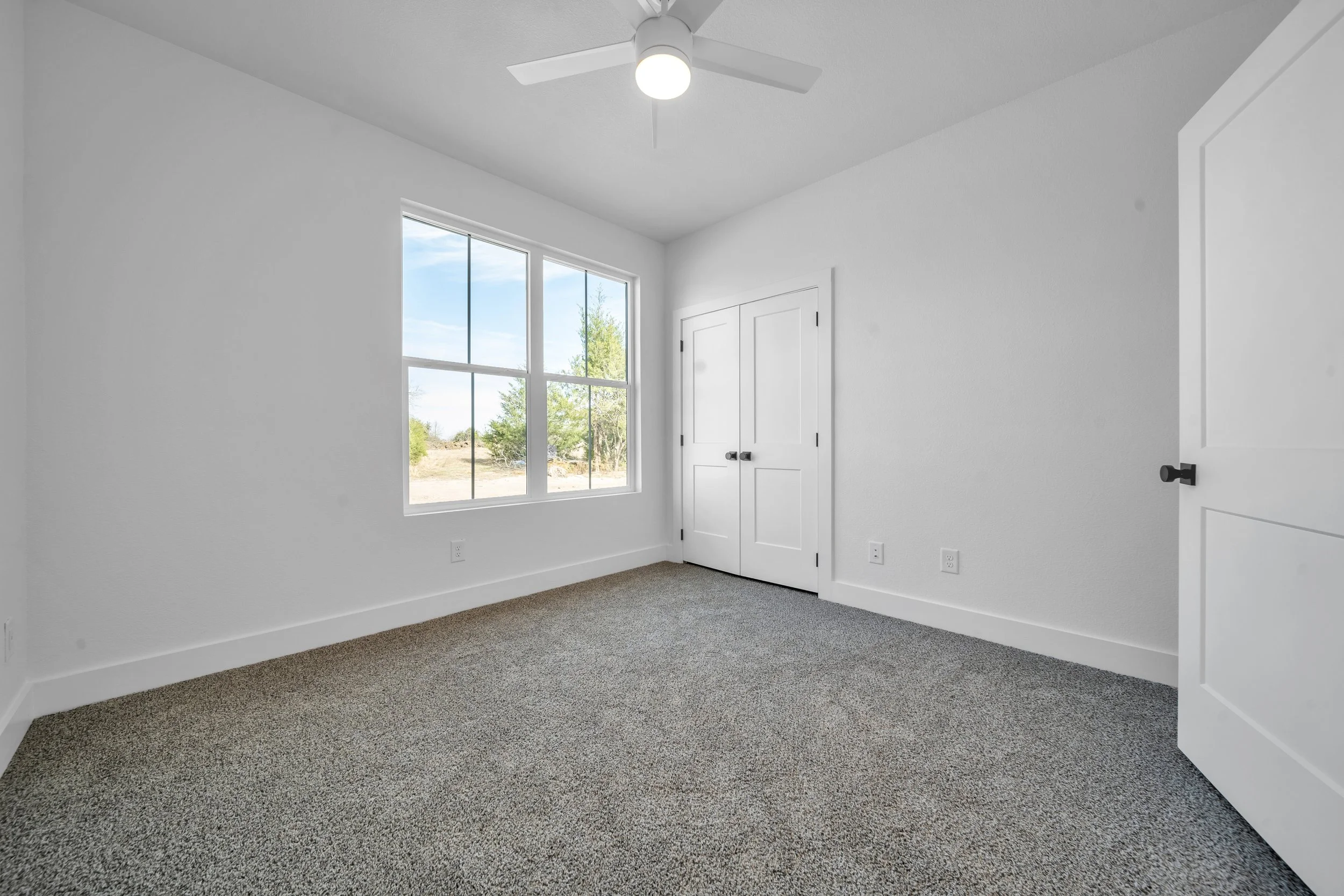 Empty bedroom with white walls, a large window, a ceiling fan, carpet flooring, and a closed door.