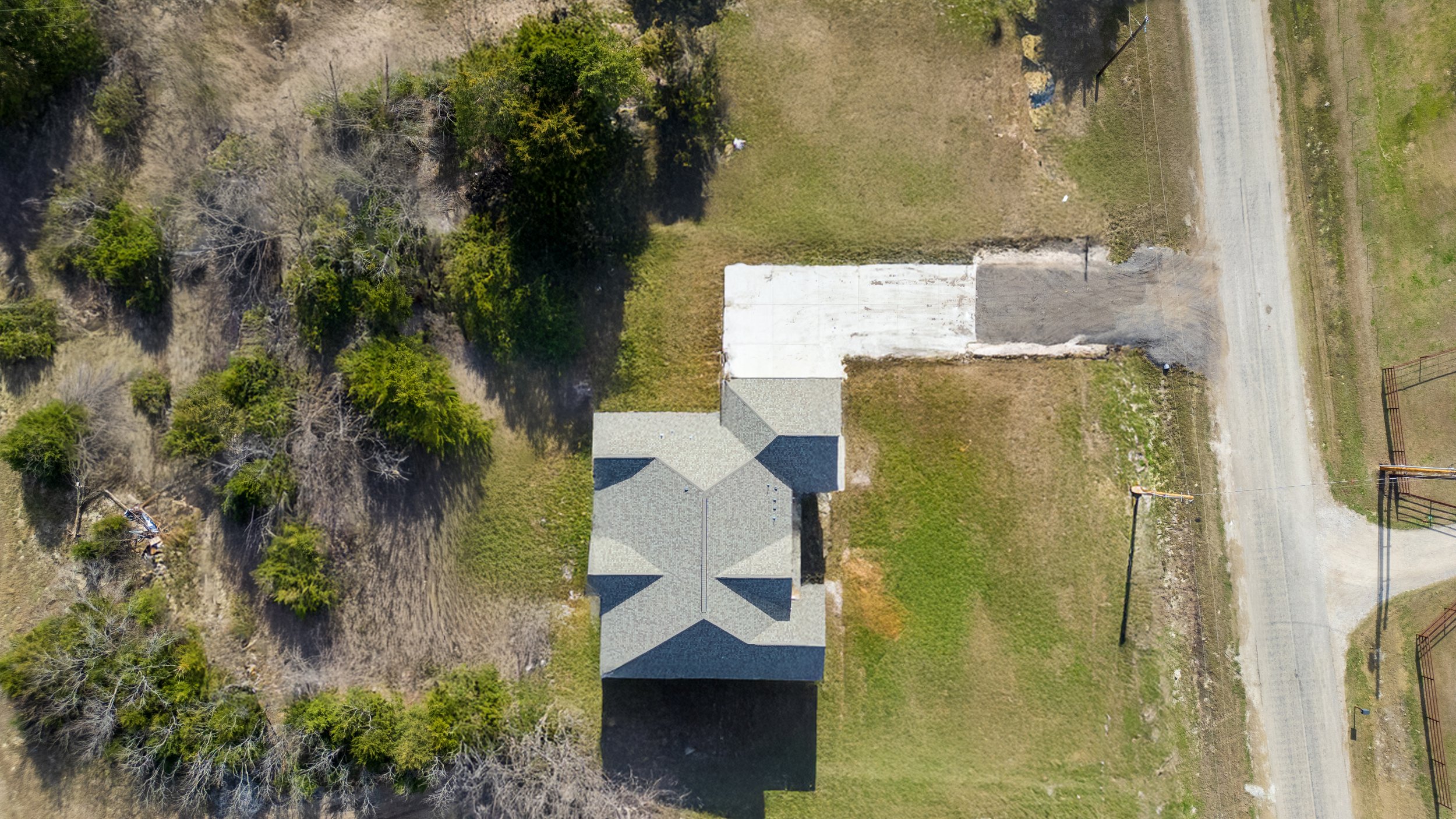 Aerial view of a house with a gray roof, surrounded by trees on the left and open grassy land on the right, with a dirt driveway leading to the street.