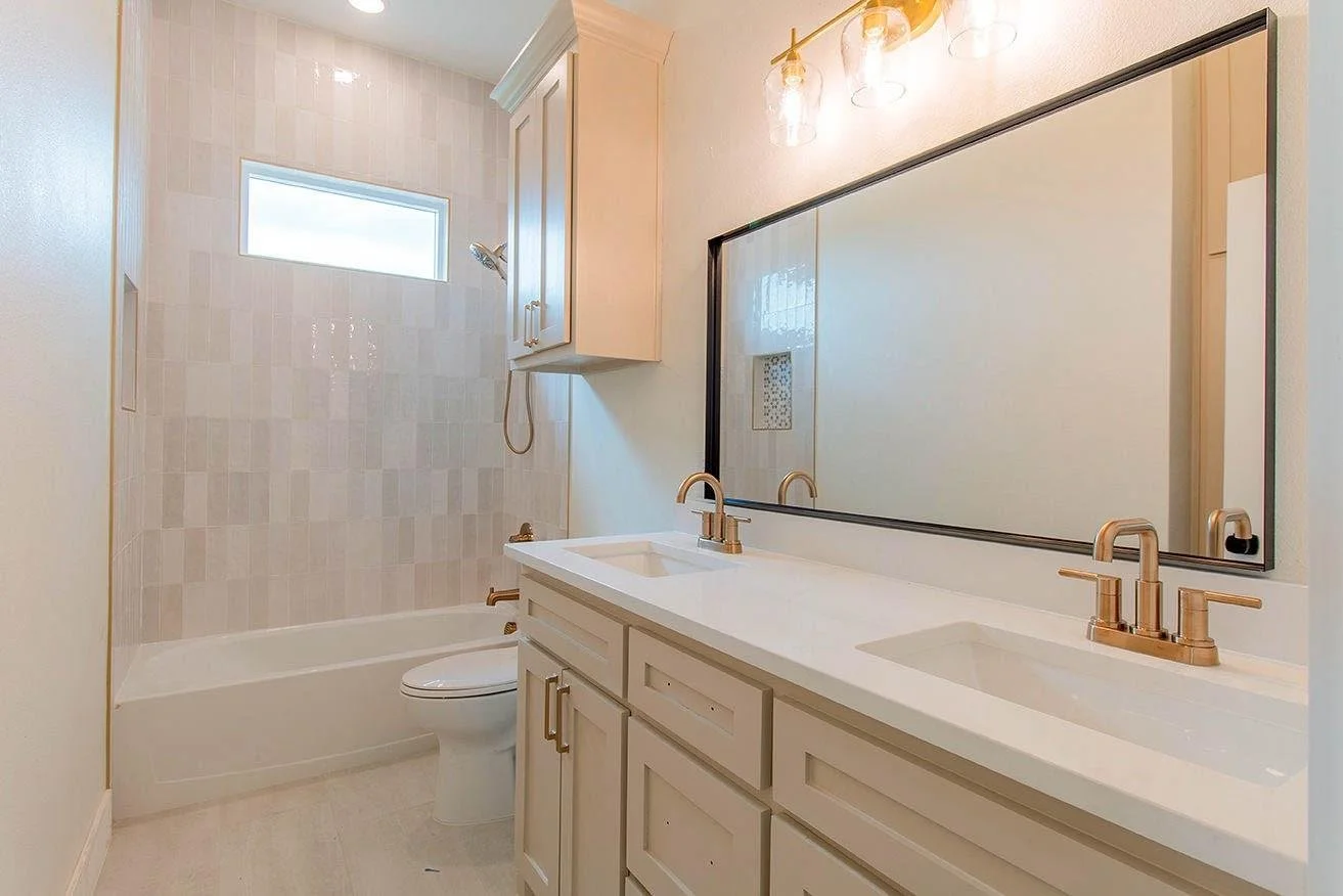 Bathroom with a double sink vanity, large mirror, beige cabinets, gold fixtures, bathtub, toilet, window, and wall-mounted cabinet.