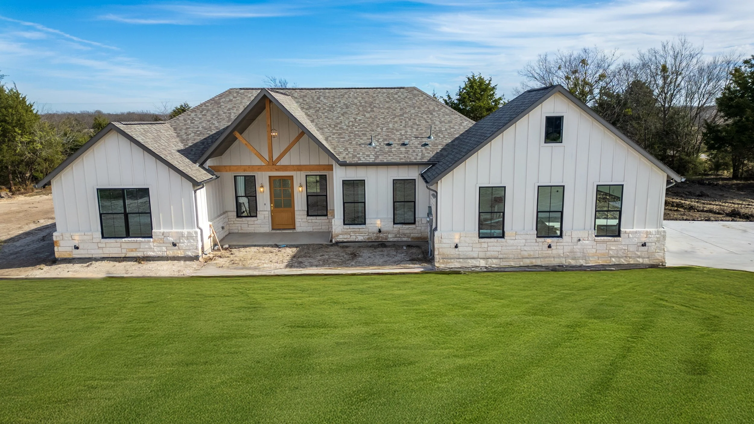 Front view of a newly constructed ranch-style house with white vertical siding, stone accents, and a gray shingled roof, surrounded by a green lawn and trees in the background.