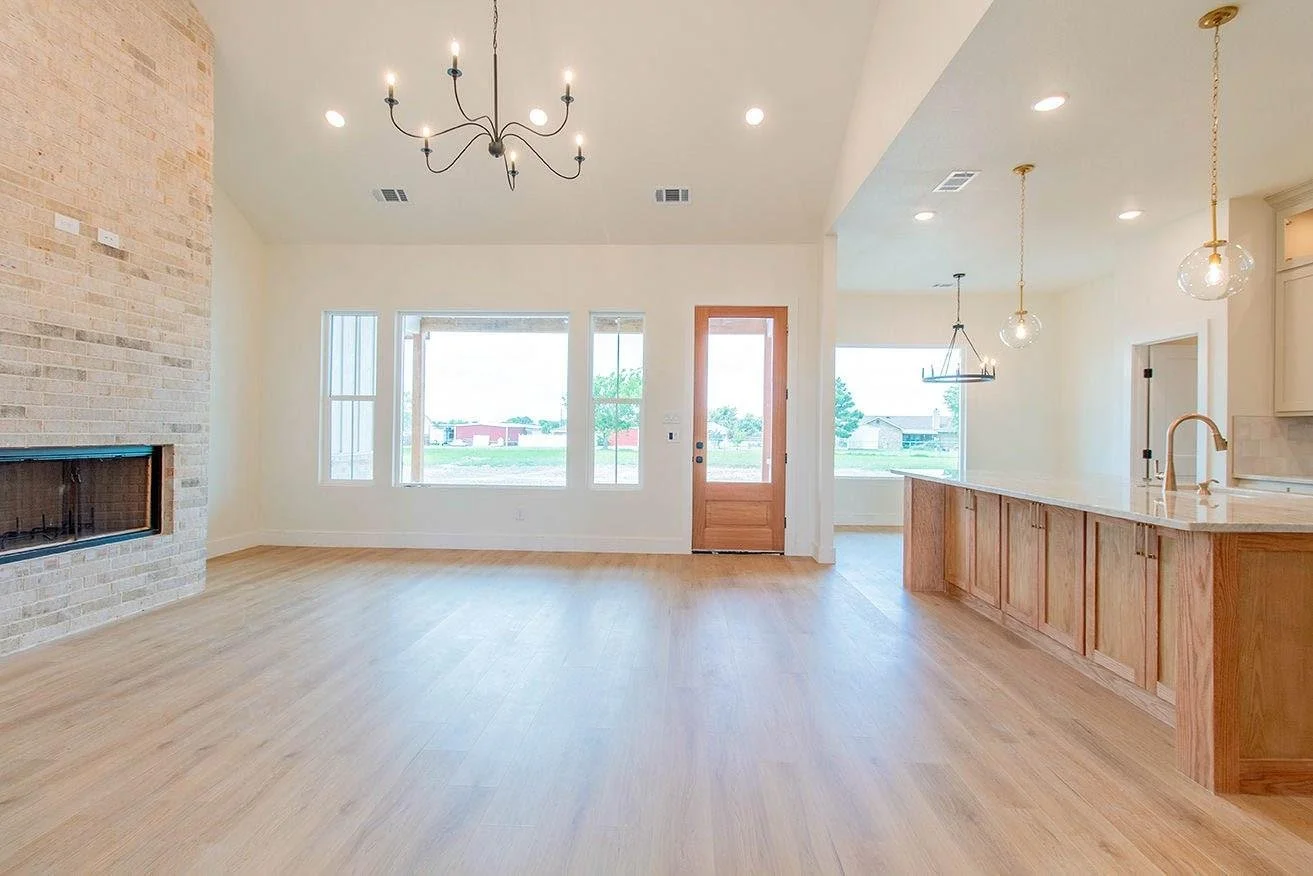 Empty living room with light wood flooring, a brick fireplace, large windows, a wooden door, and a kitchen bar with pendant lights.