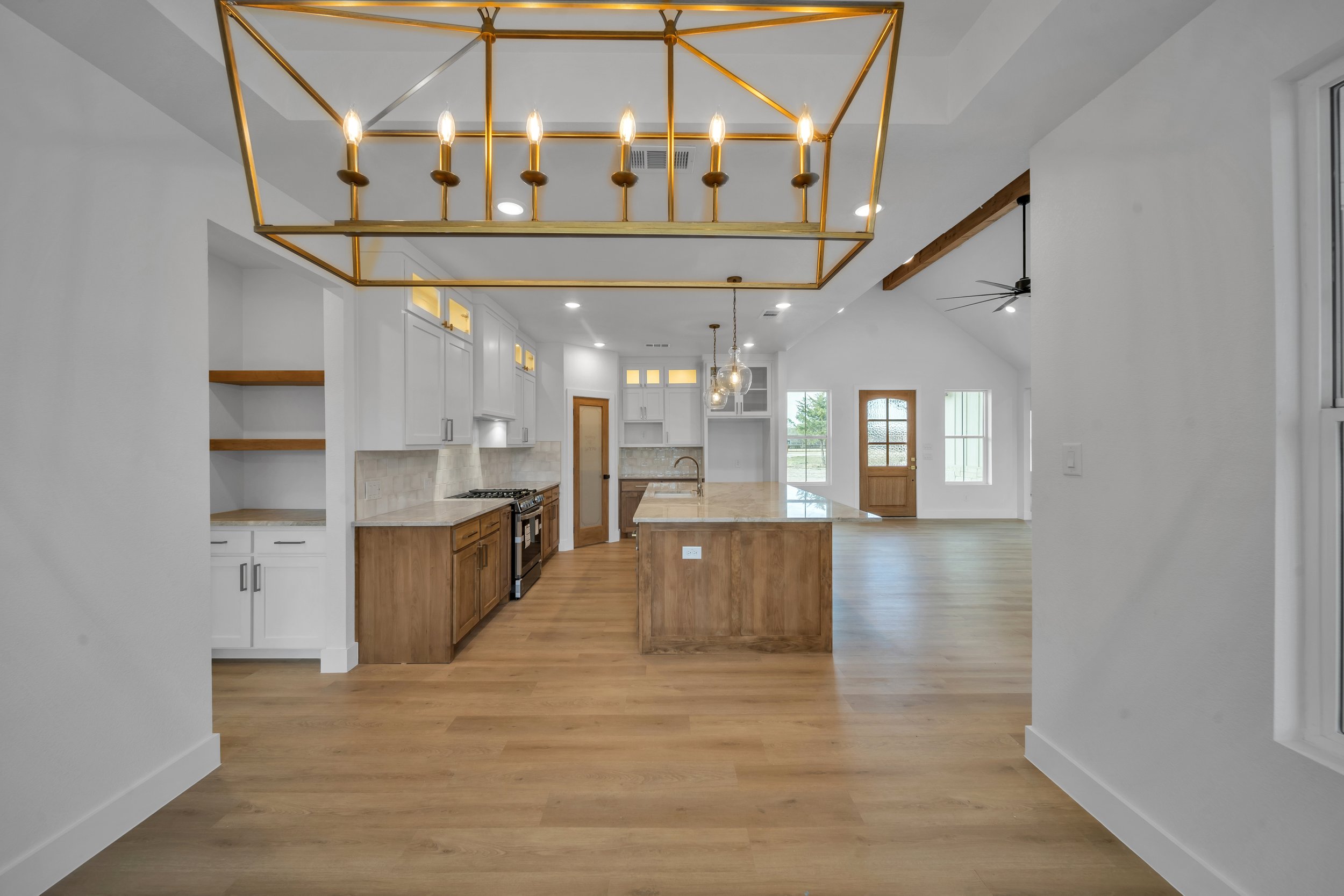 Open-concept kitchen with wooden cabinets and an island, white upper cabinets, light wood flooring, gold chandelier over the island, pendant lights, and a view into a bright living area with large windows.