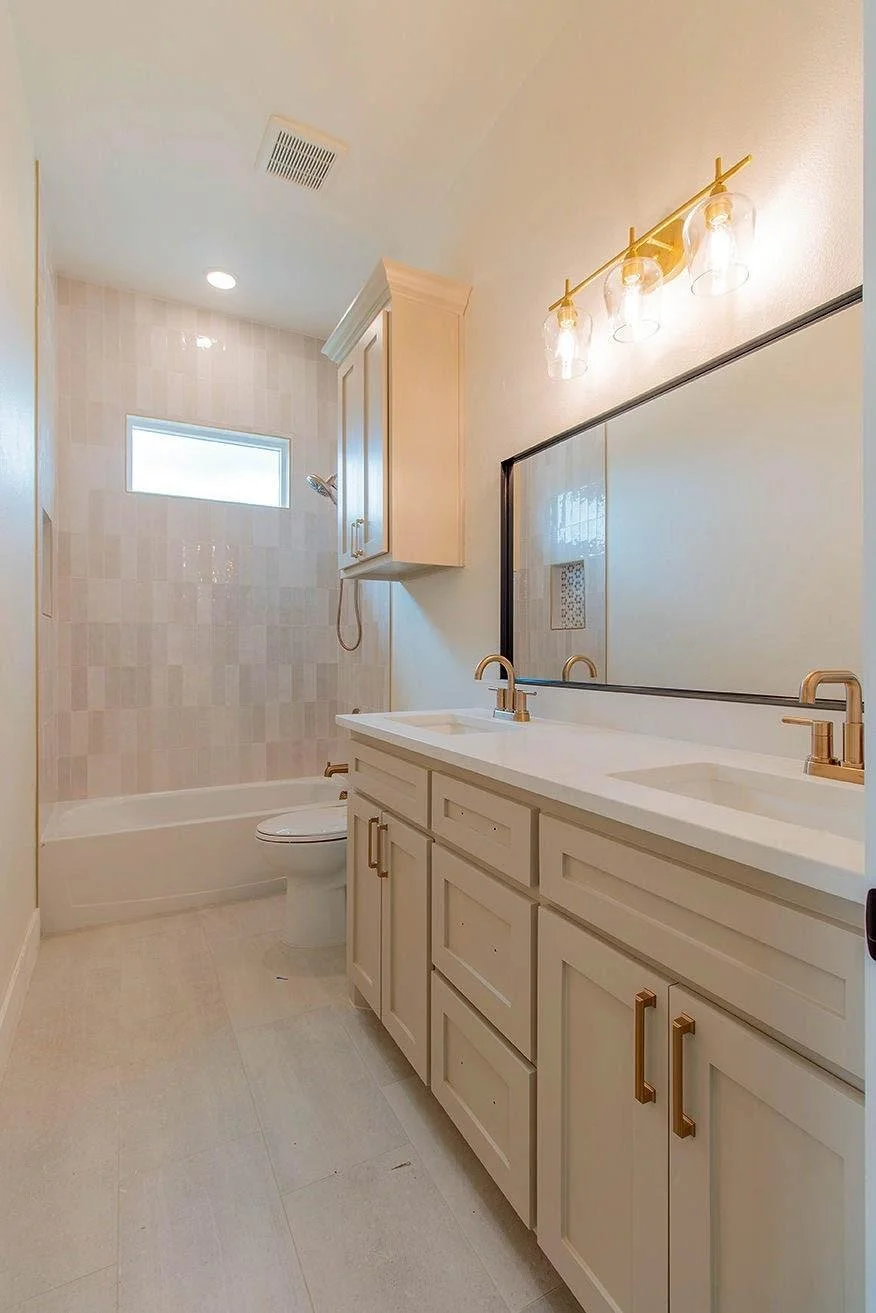 Modern bathroom with double vanity, large mirror, gold fixtures, white cabinetry, walk-in shower with small window, and beige tiled walls and floor.
