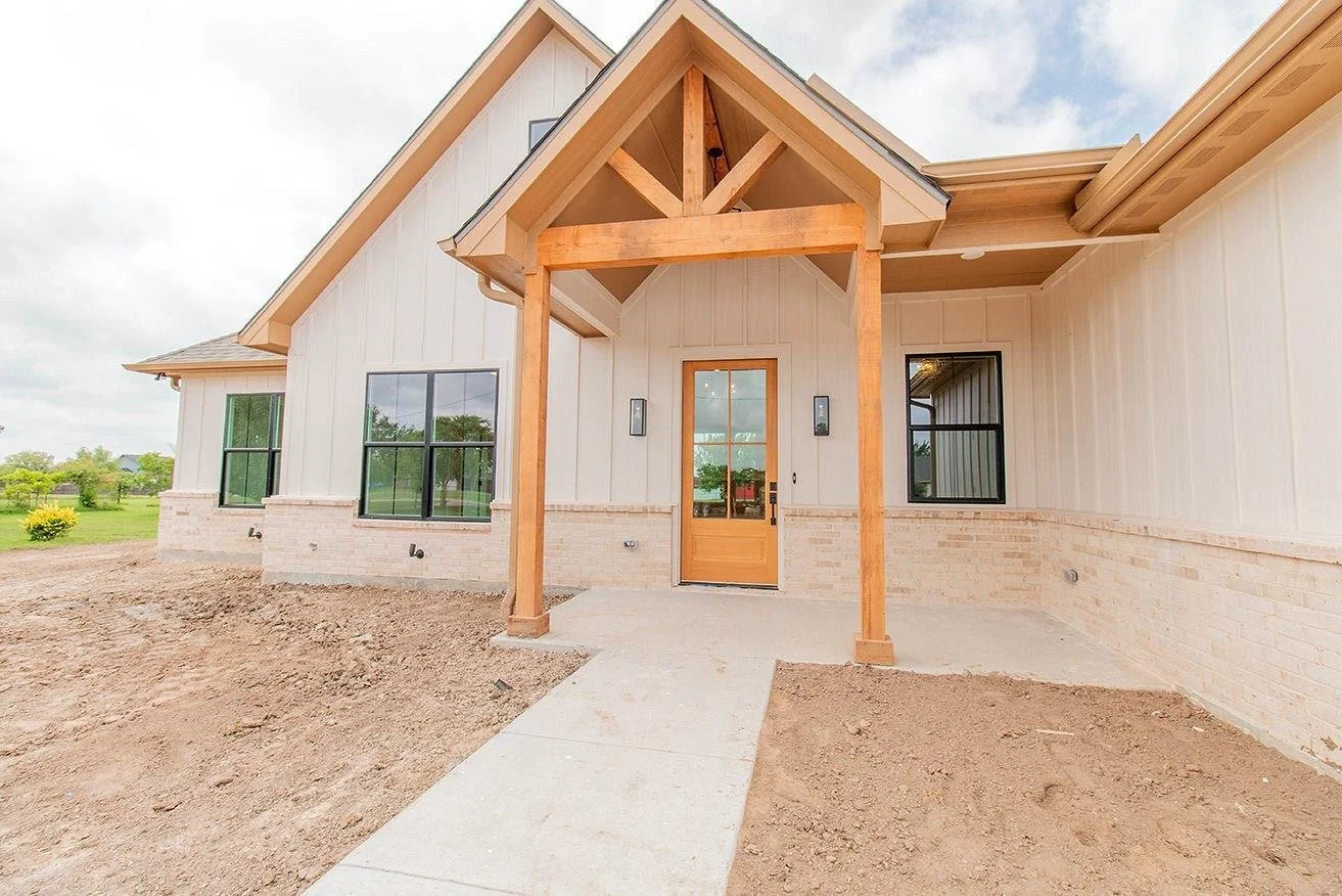 Newly constructed house with a brick exterior, multiple large windows, and a wooden front porch with a gable roof, surrounded by dirt and grass under a cloudy sky.