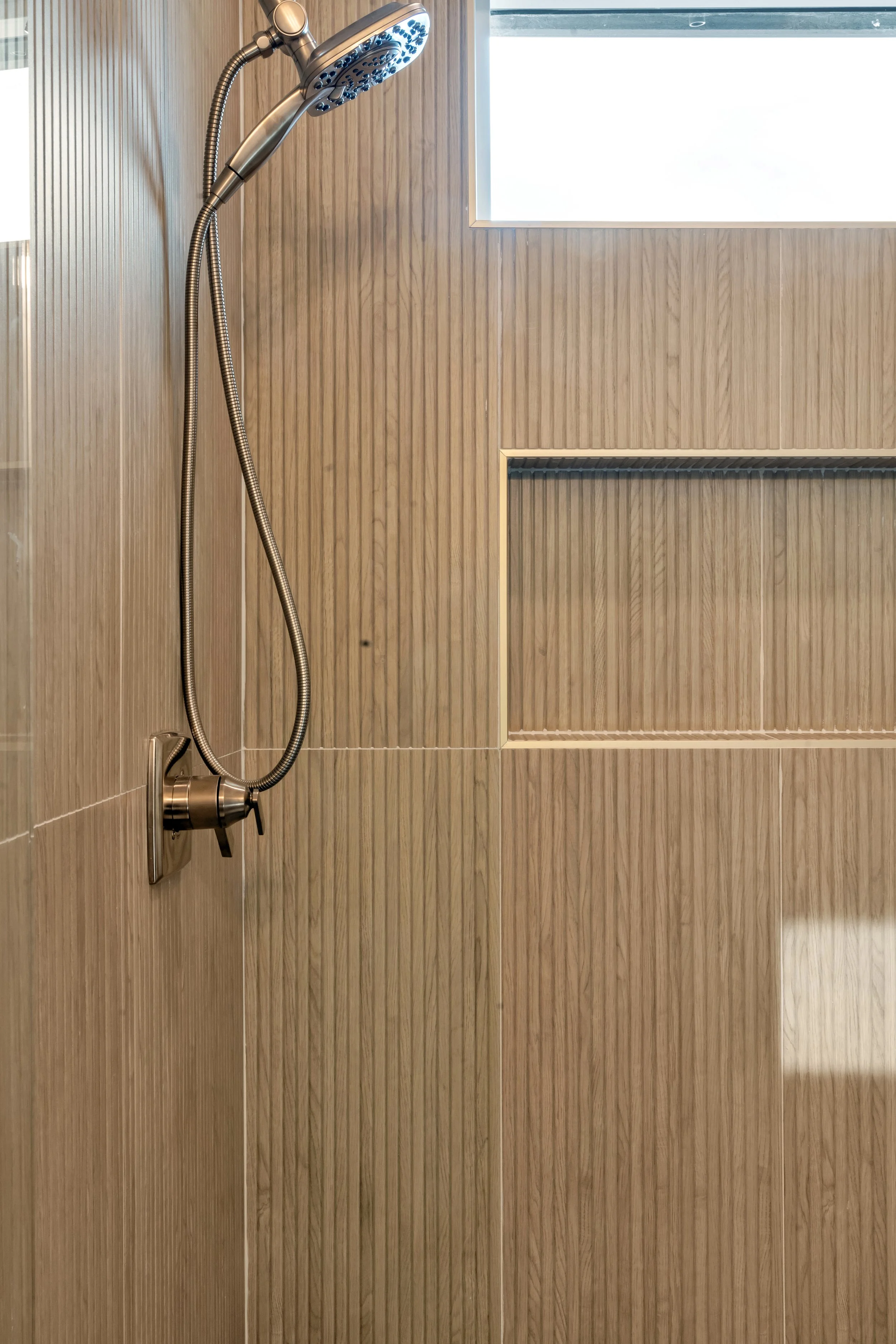 Shower with a chrome handheld showerhead mounted on a wood-textured beige wall, with a small window above.