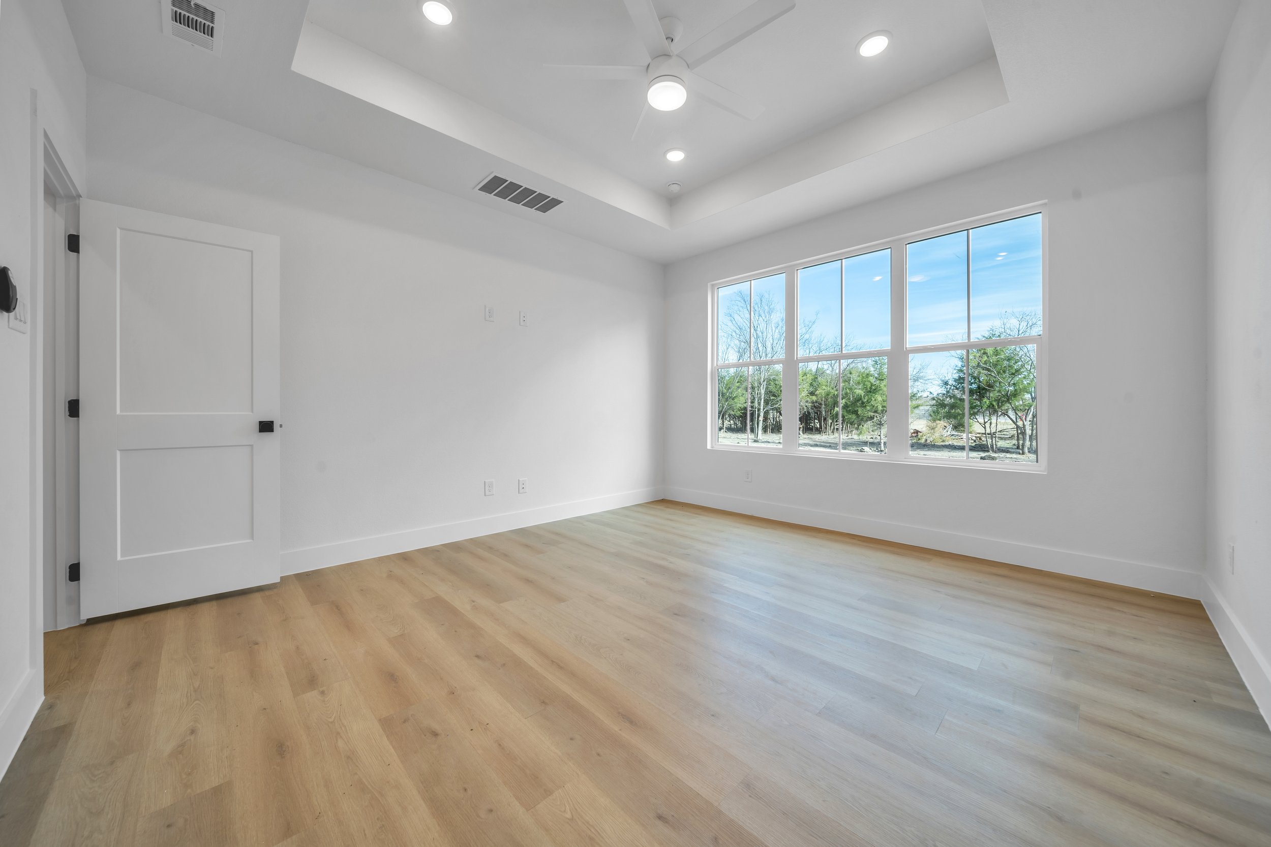 Empty room with white walls, a large window showing trees outside, a ceiling fan, and light wood flooring.