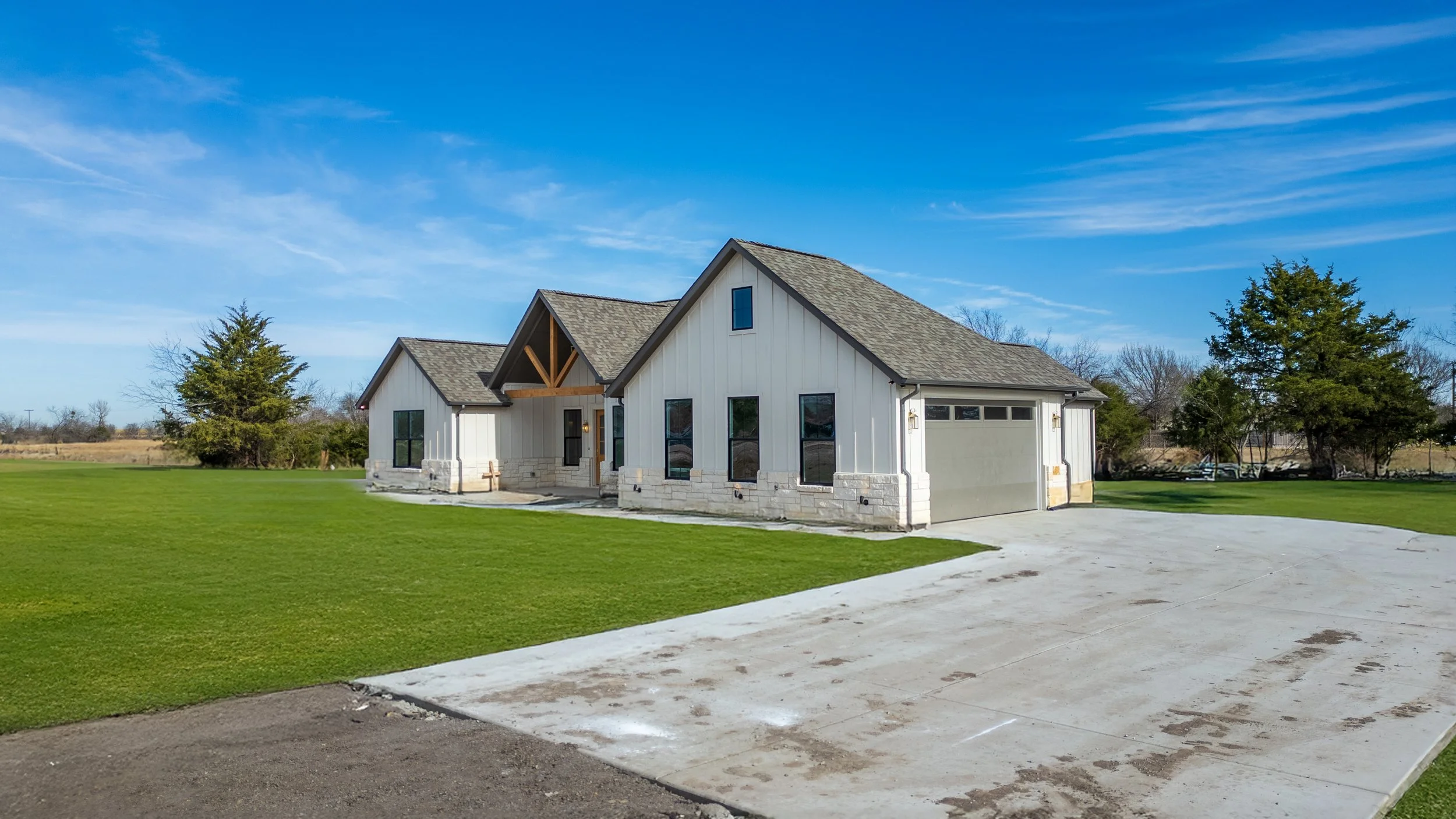 A modern house with white vertical siding, stone accents at the base, and a gray shingle roof, situated on a large grassy lot under a clear blue sky.
