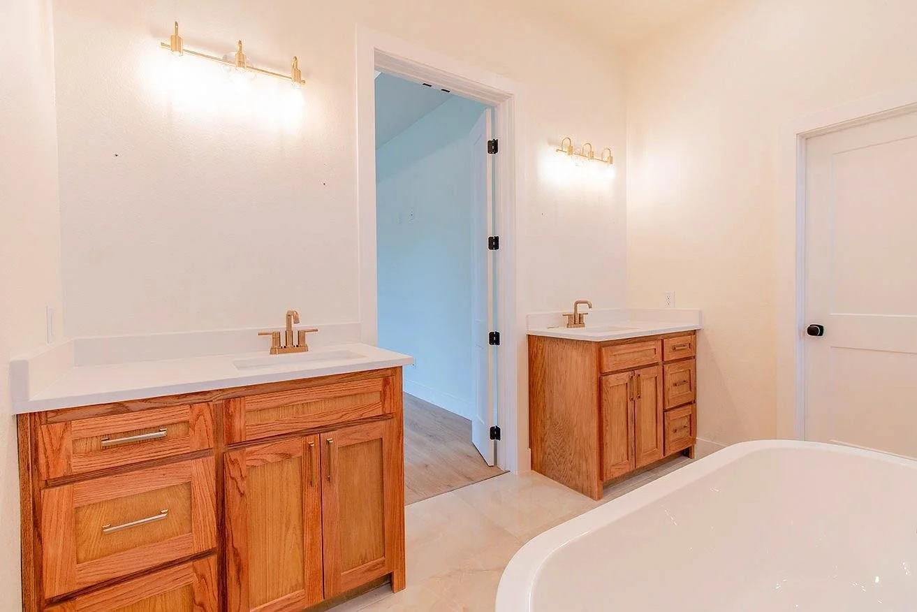 A bathroom with two wooden vanities with white countertops, golden faucets, and wall-mounted light fixtures, a partial view of a bathtub, and the door to a hallway.