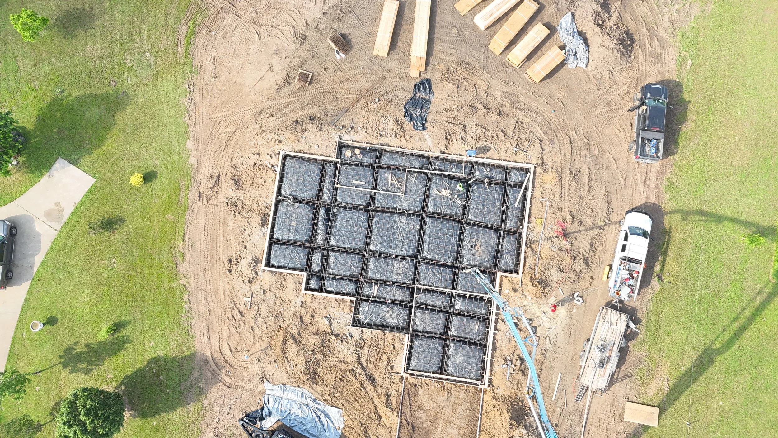 Aerial view of a construction site showing the foundation and framework of a building with steel reinforcement bars and black plastic sheeting, surrounded by dirt and grass, with construction vehicles and materials nearby.