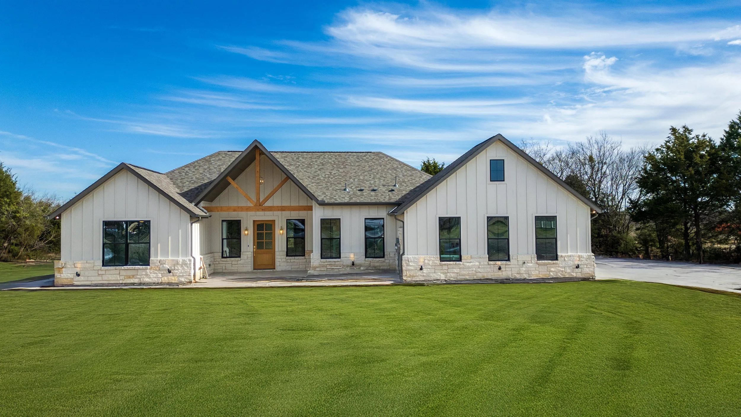 Newly constructed modern farmhouse with white exterior, black window frames, exposed wooden beams, and a stone foundation, surrounded by a green lawn and trees under a partly cloudy sky.