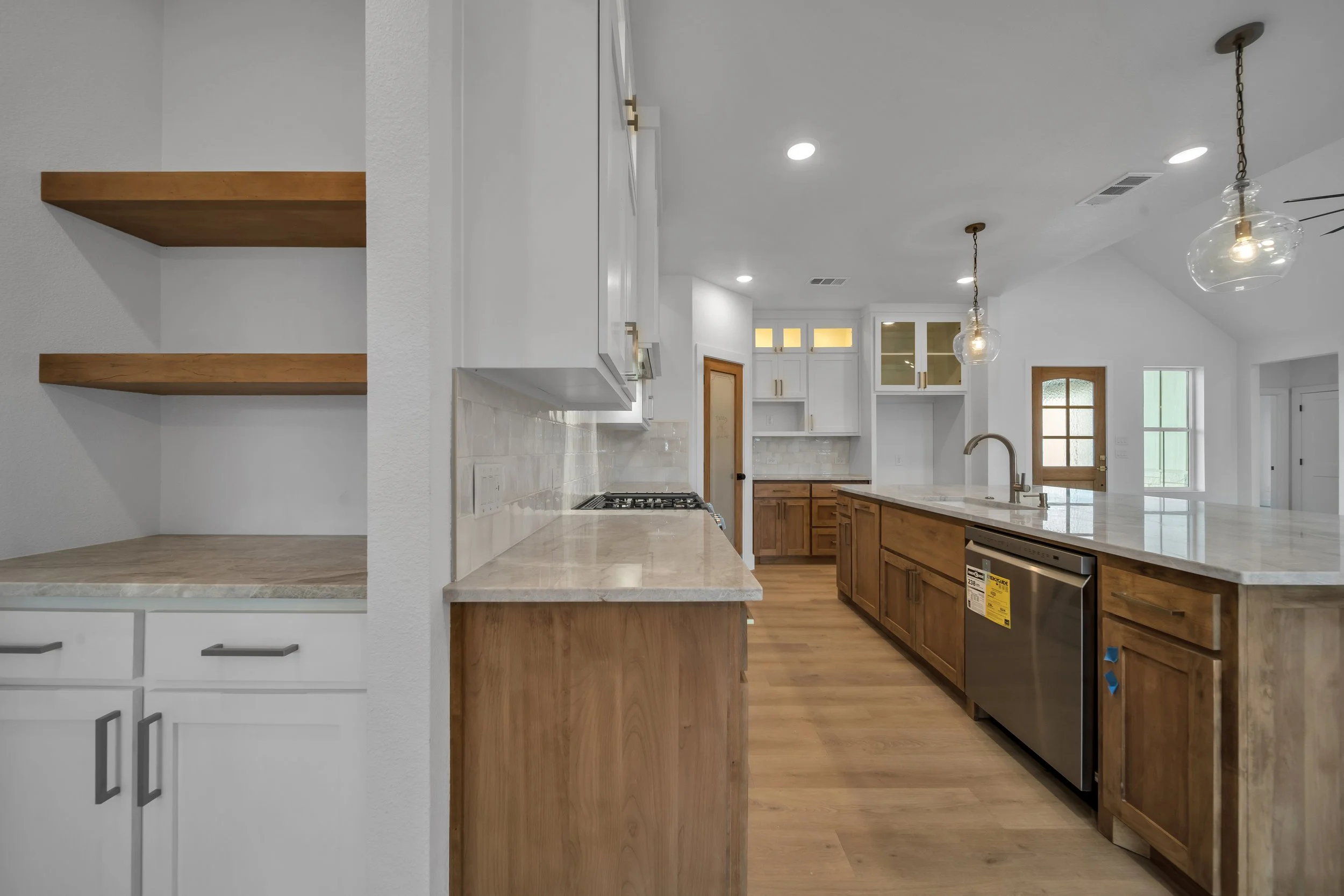 Modern kitchen with white and wooden cabinets, marble countertops, hardwood flooring, pendant lights, and a view into a living area with windows.