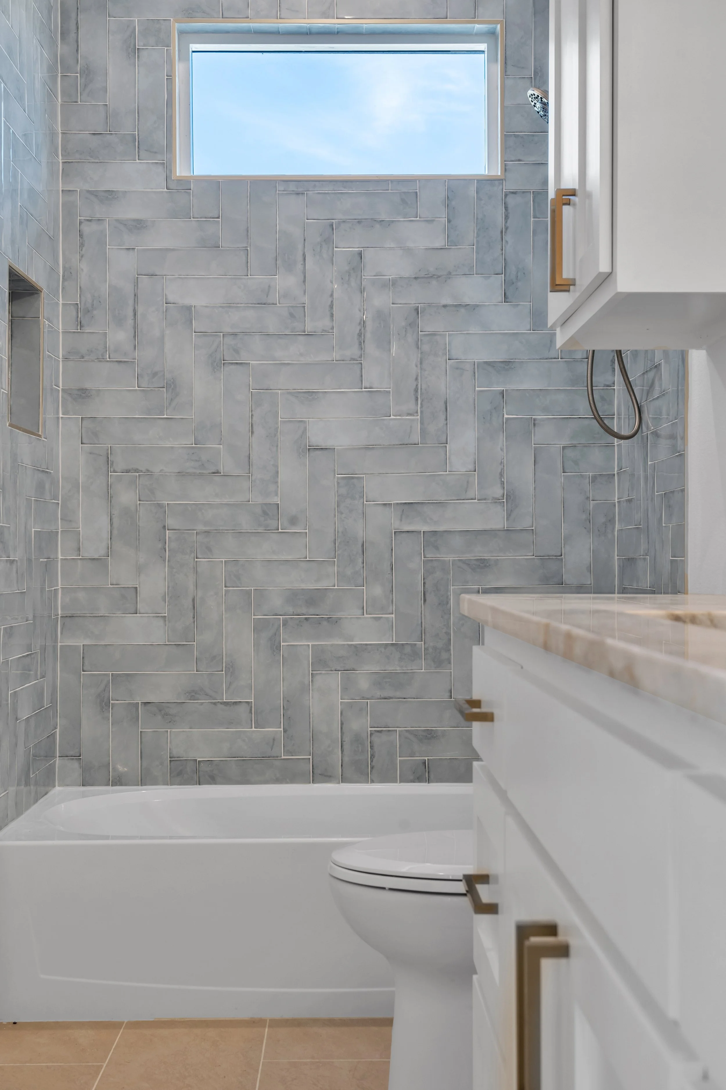A modern bathroom with a bathtub, a toilet, gray tiled walls with a herringbone pattern, a window letting in natural light, and a white vanity with gold hardware.