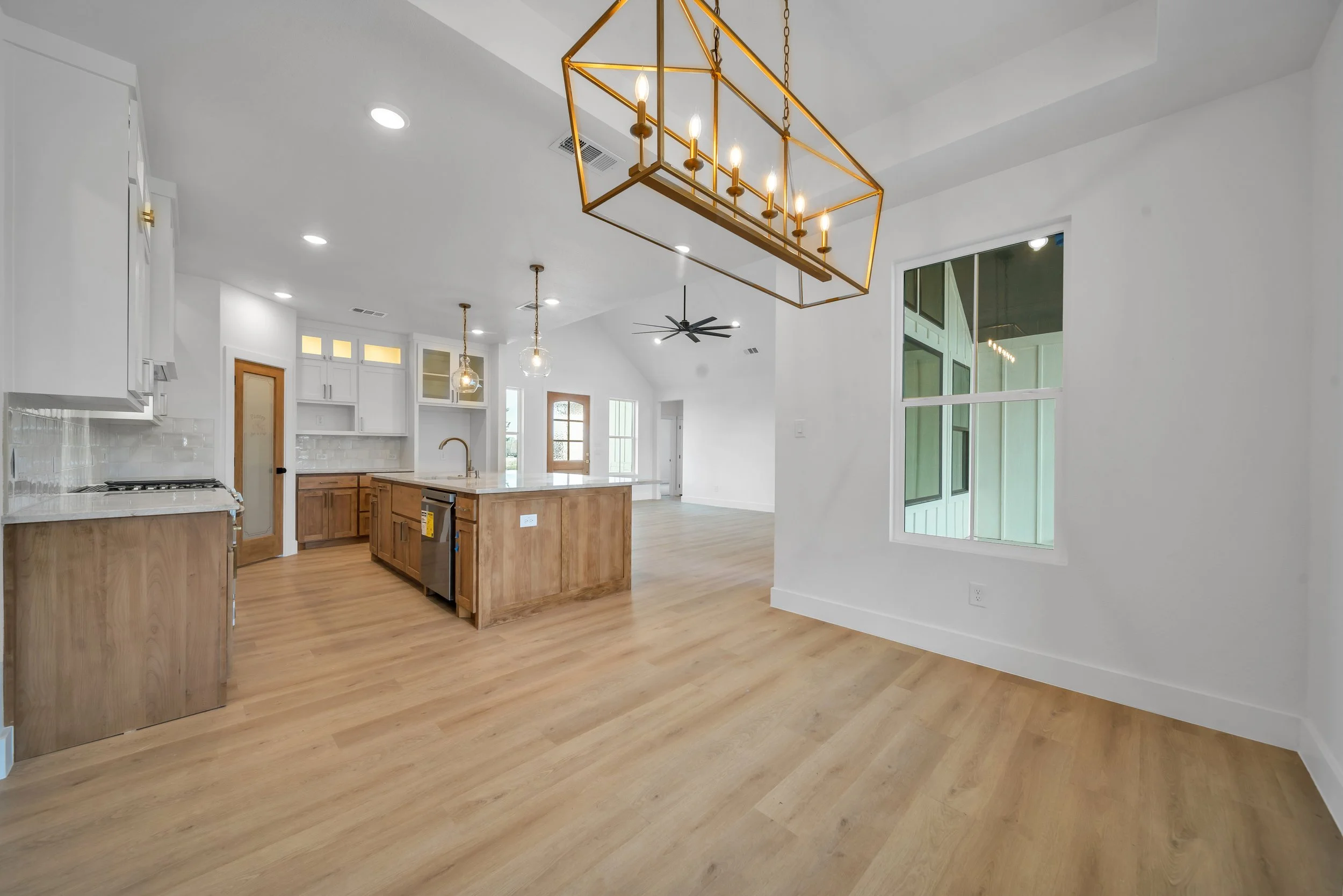 Open-concept kitchen and living space with wooden flooring, white walls, and large windows. Kitchen features an island with a sink, white cabinets, and a tile backsplash. A gold chandelier hangs over the dining area, and pendant lights hang above the