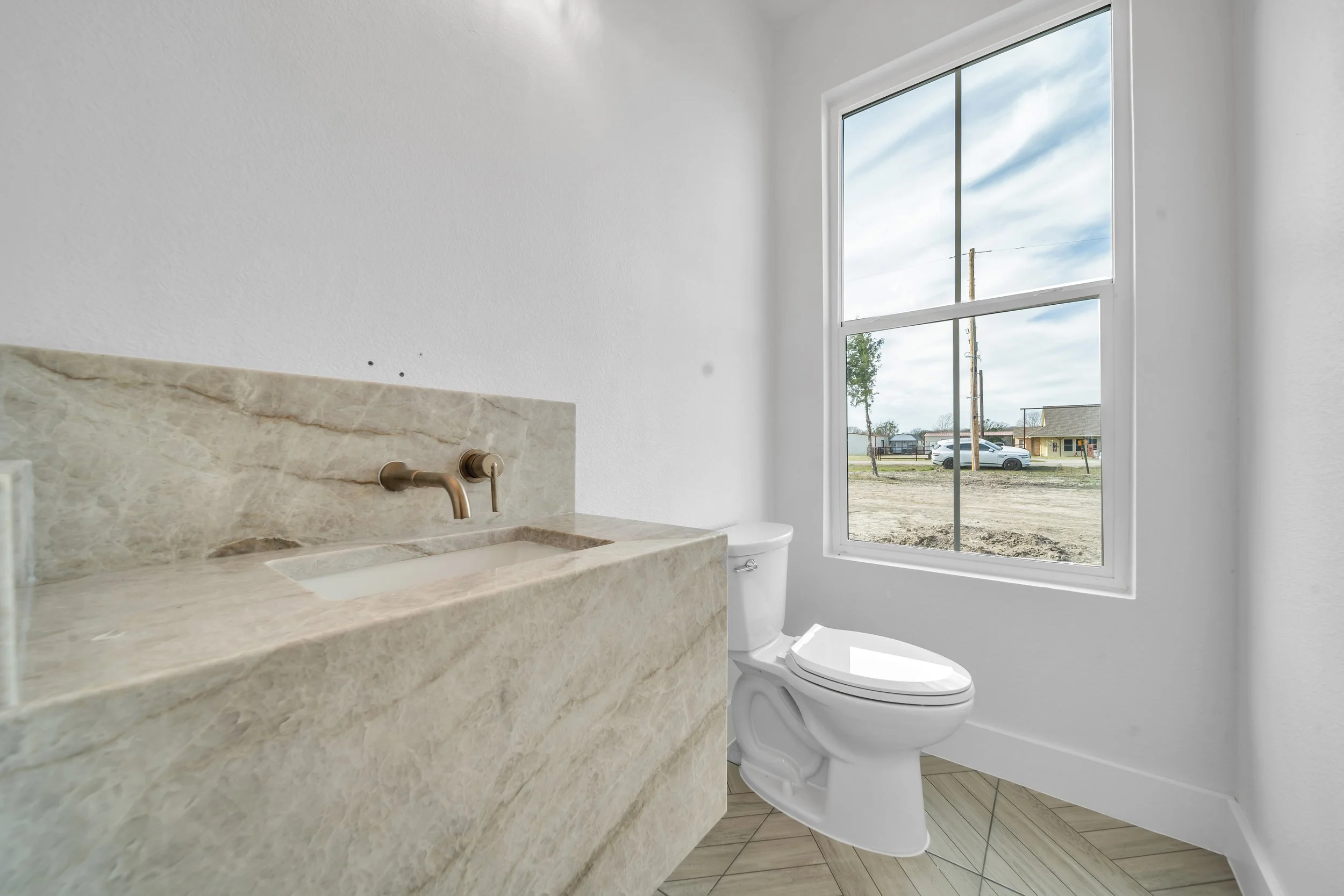 Interior of a bathroom with a white toilet and a beige marble sink, next to a large window showing a view of a street with parked cars and power lines, and a partly cloudy sky.