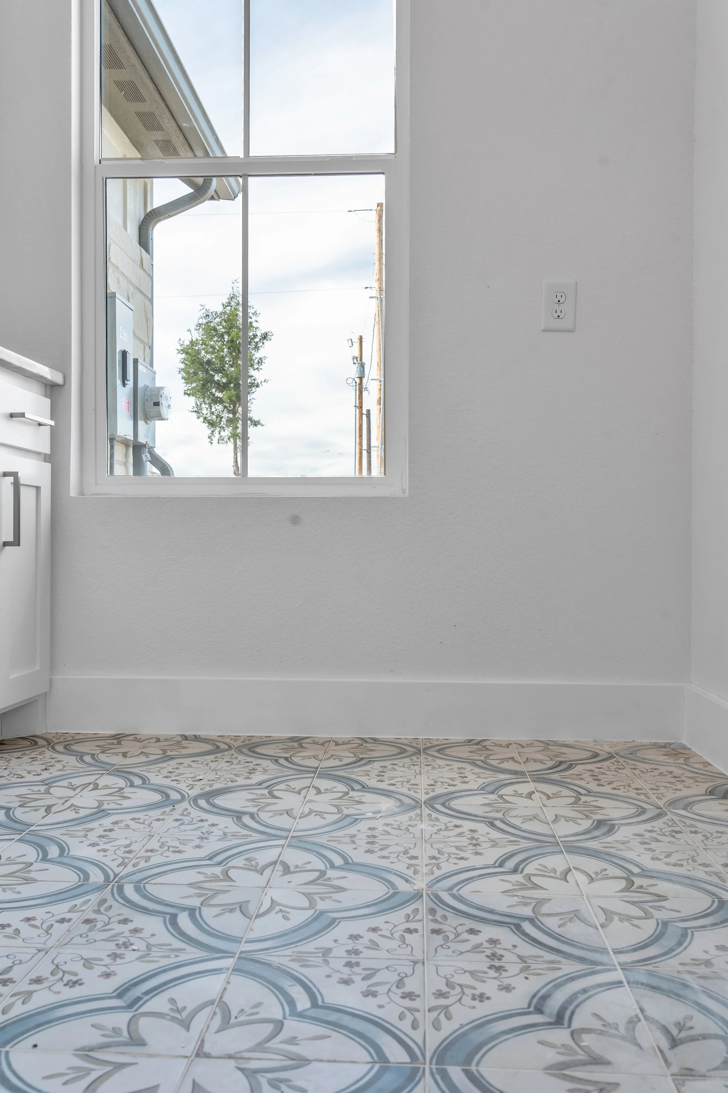 Empty room with decorative patterned ceramic tile flooring, white walls, a large window showing an outdoor view with utility poles and a blue sky, and a white electrical outlet on the wall.