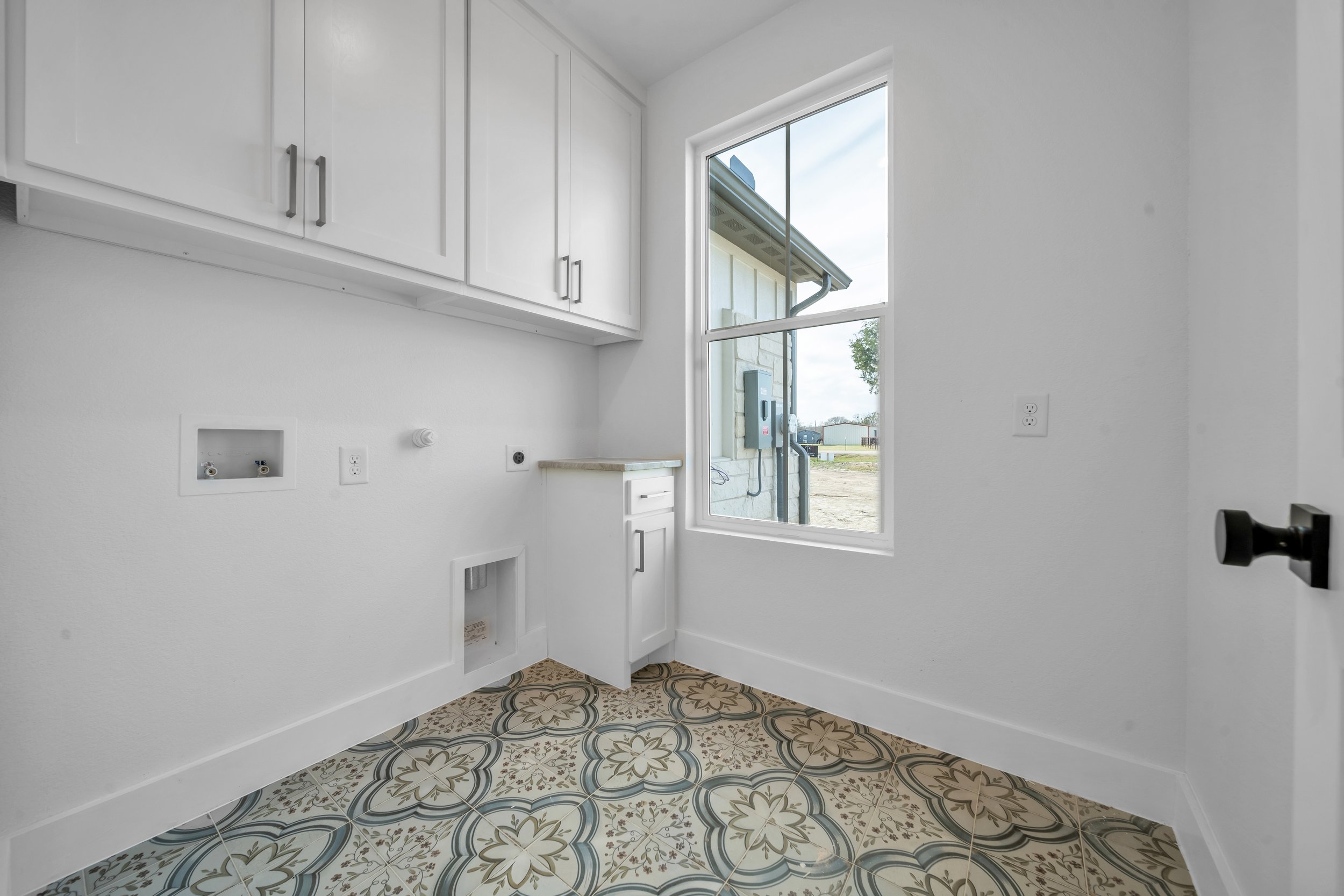 Empty laundry room with white cabinets, a window, electrical outlets, and colorful patterned tile flooring.