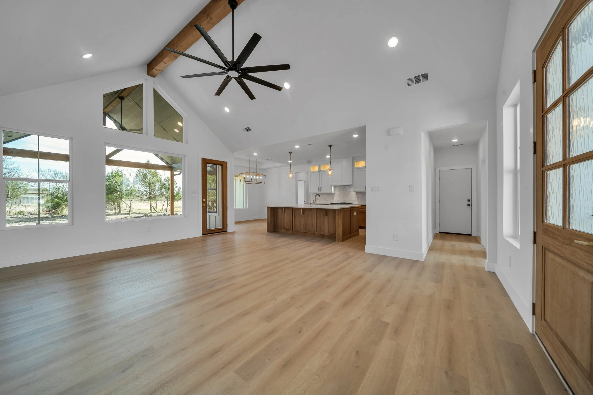 Empty modern living room with wood floors, large windows, a black ceiling fan, and an open kitchen with an island, pendant lights, and white walls.