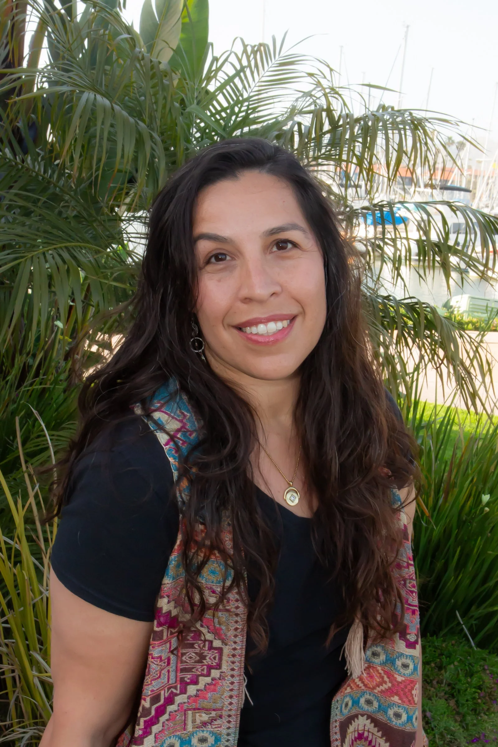 A woman with long, wavy dark hair and a radiant smile, wearing a black top, a gold necklace with a pendant, and a colorful patterned shawl, standing outdoors with lush green plants and a marina with boats in the background.