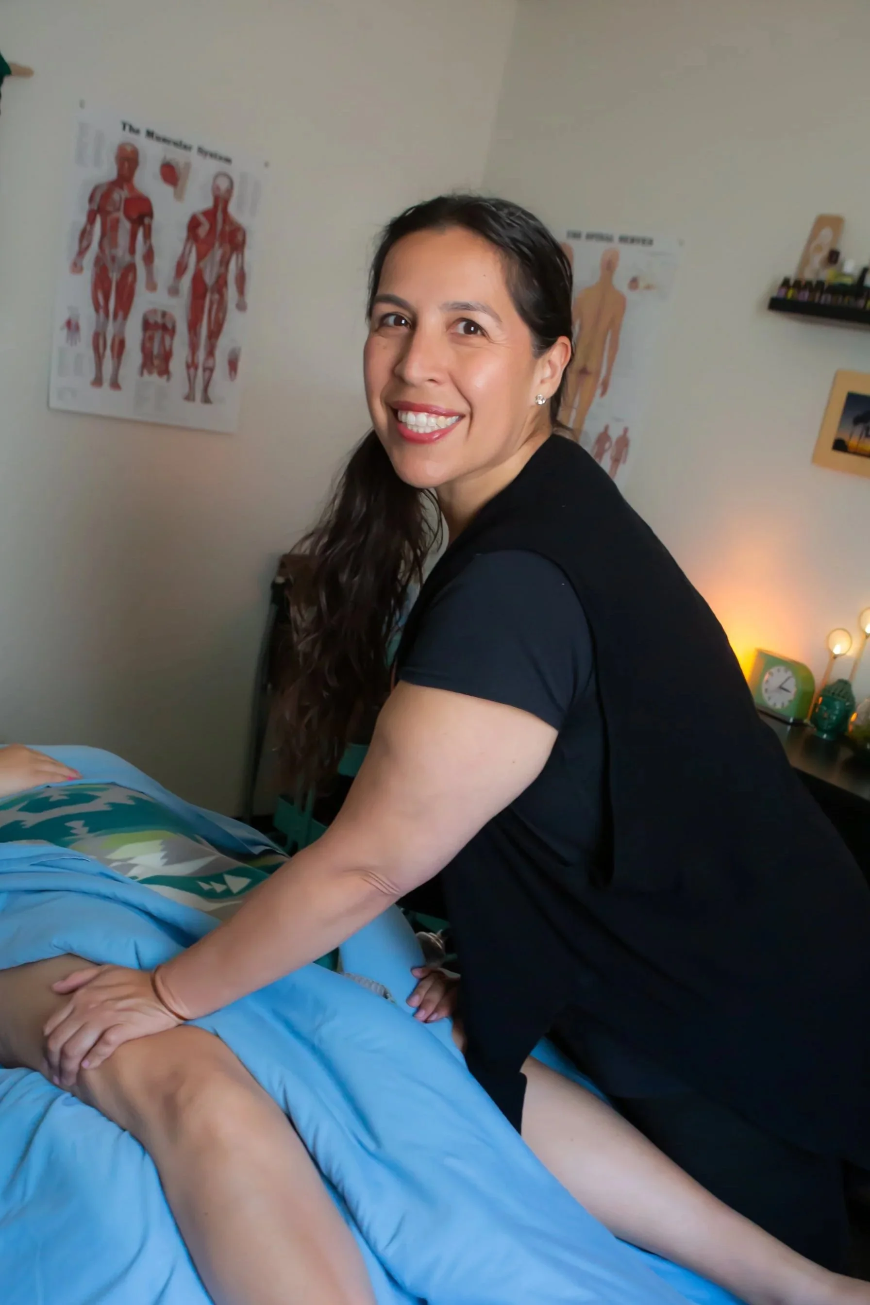 A woman giving a massage to a person lying face down on a massage table with a blue blanket. The woman is smiling at the camera, wearing a black shirt, with posters of human anatomy on the wall behind her.