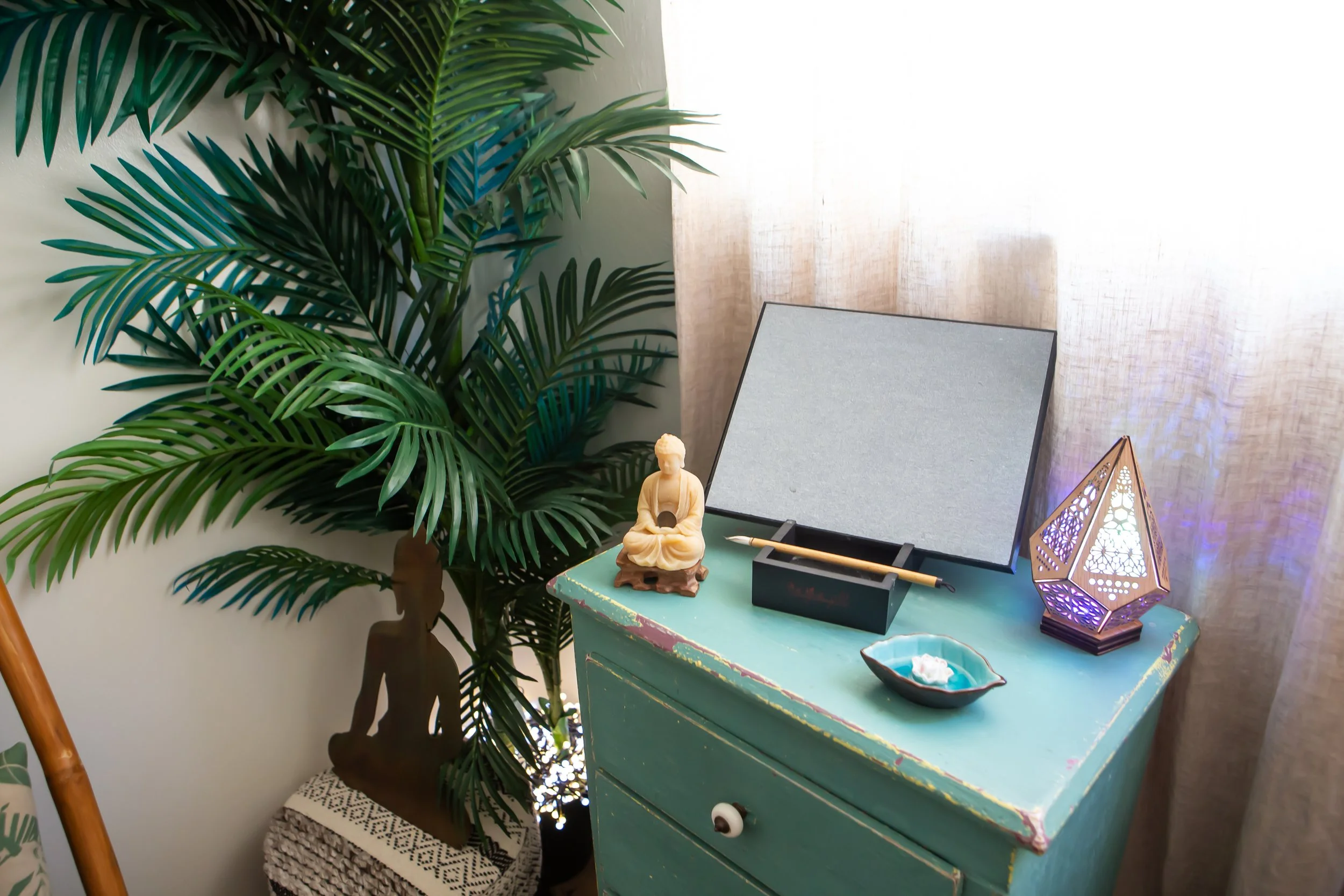 A blue wooden table with a decorative figurine, a black pen holder, and a colorful lamp. Behind the table is a large leafy houseplant and a light curtain in the background.