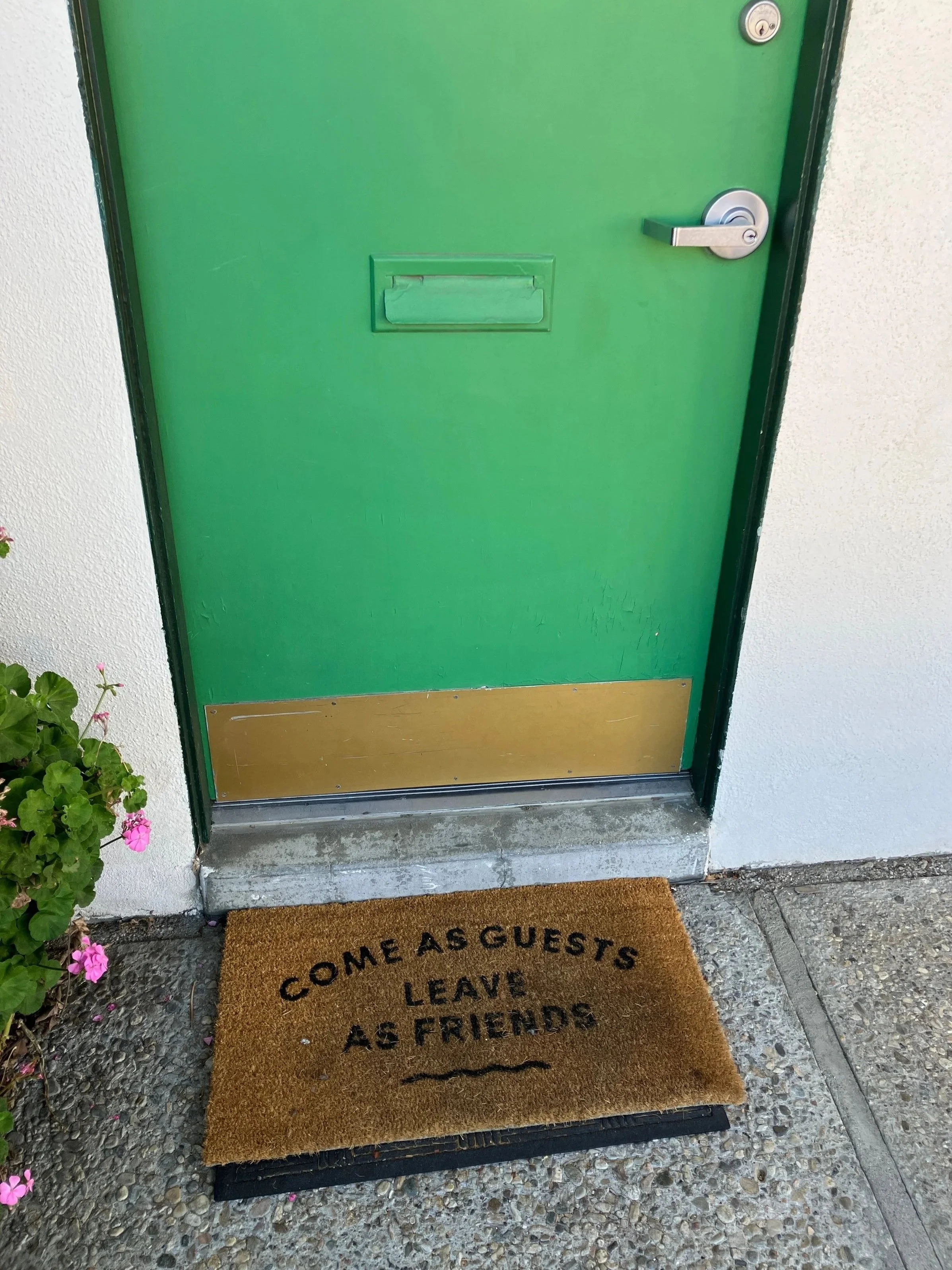Green door with a mail slot and door handle, white wall on the right, pink flowers to the left, and a brown doormat on the ground that reads "Come as guests, leave as friends"