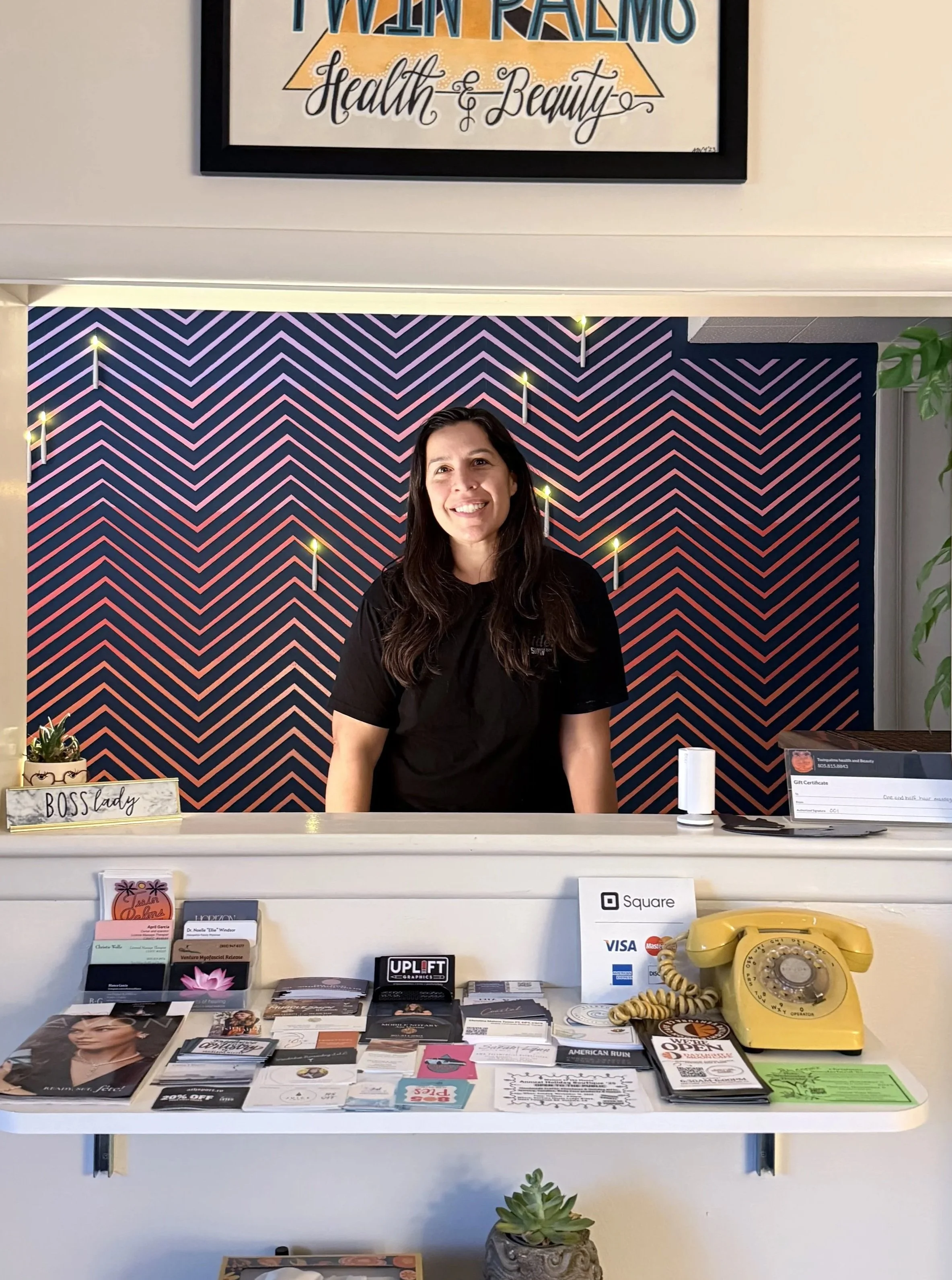 A woman smiling behind a reception counter with various brochures, flyers, and business cards on display, a yellow rotary telephone, and a small green succulent plant in a decorative pot in the foreground. Behind her is a colorful geometric patterned wall with small lights, and a sign with her title, 'Boss Lady.'