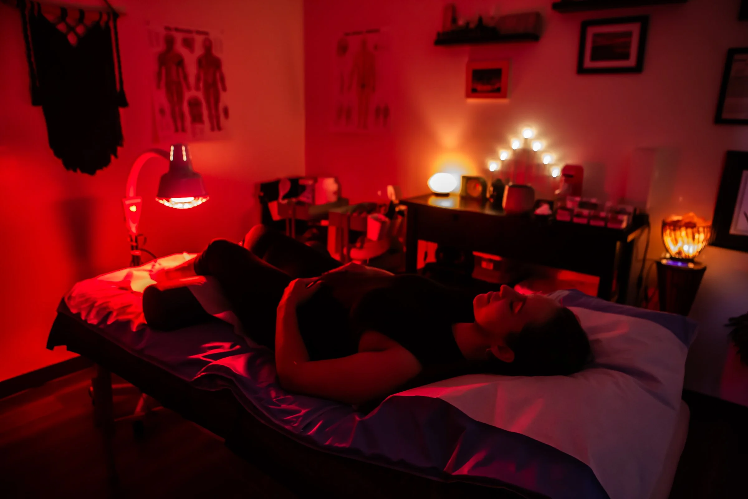 A person lying on a massage table in a dimly lit room illuminated with red and warm lighting, with medical posters and framed pictures on the walls, and various small lamps and objects on a desk and nightstand.
