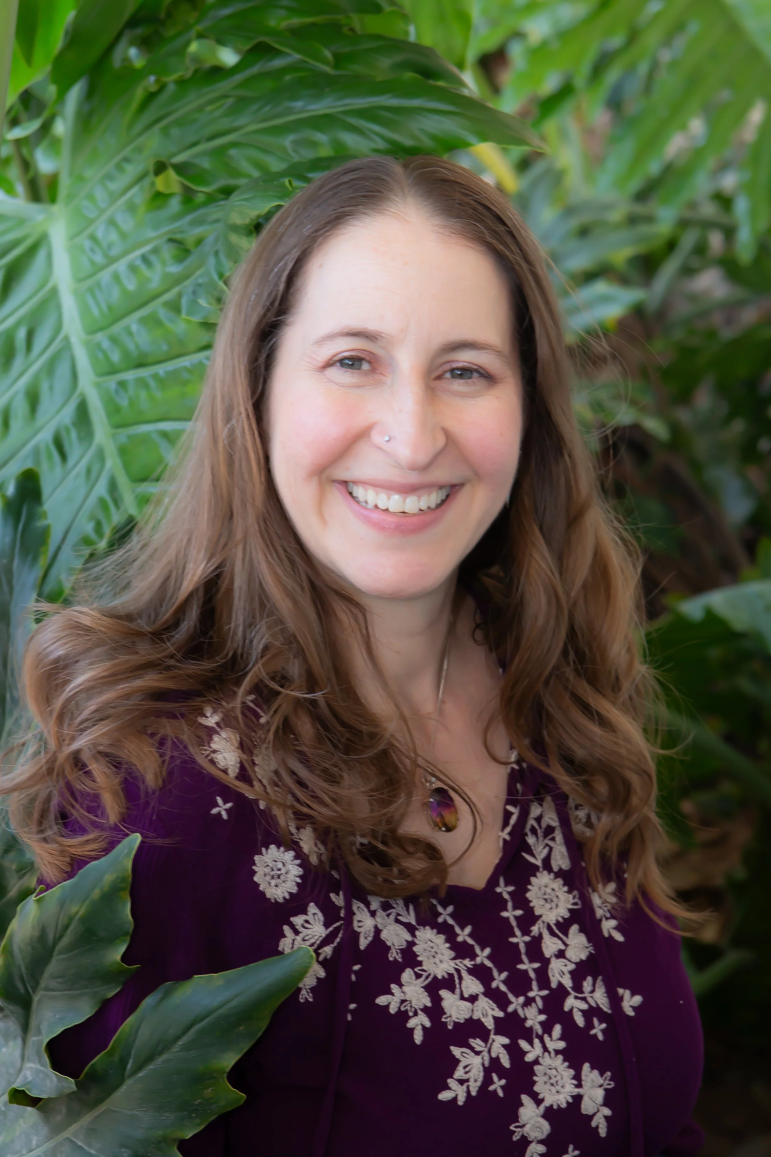 A woman with long wavy brown hair, smiling, wearing a purple top with white floral embroidery, standing among large green leaves.