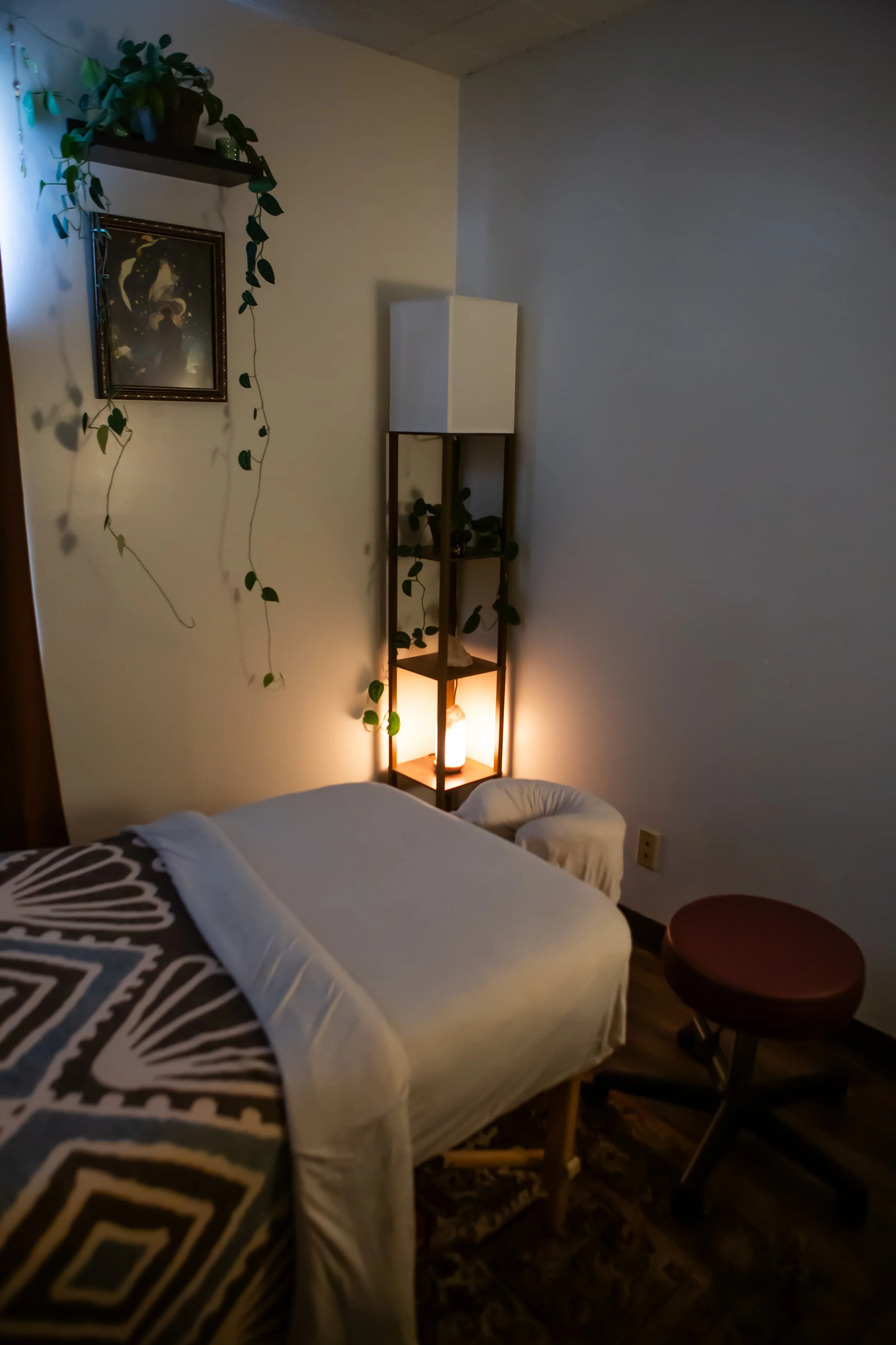 A treatment room with a massage table, a small round red stool, a wooden shelf with potted plants and a lamp, a wall hanging, and a corner with a white cabinet.