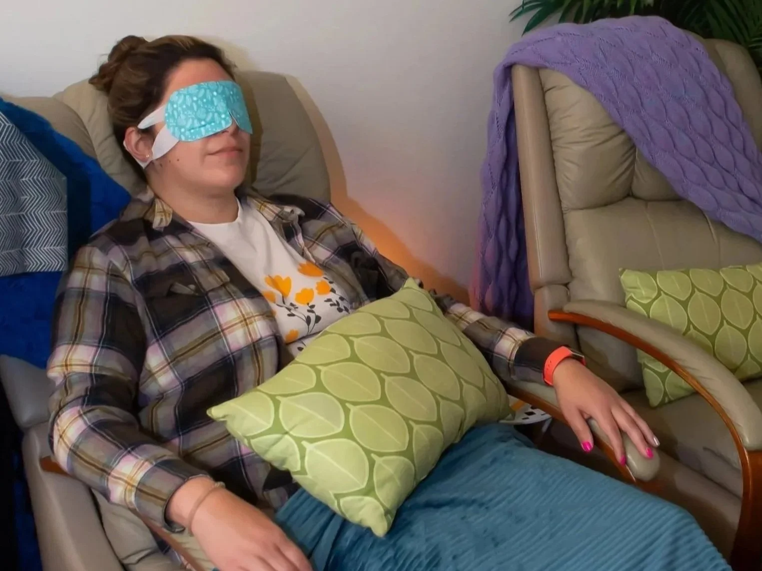 Woman sitting in a recliner chair with a blue sleep mask over her eyes, wearing a plaid shirt and holding a green pillow, in a relaxing setting.