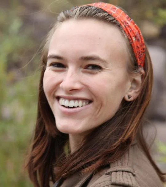 Close-up of a smiling young woman with long brown hair, wearing an orange headband, outdoors in a natural setting.
