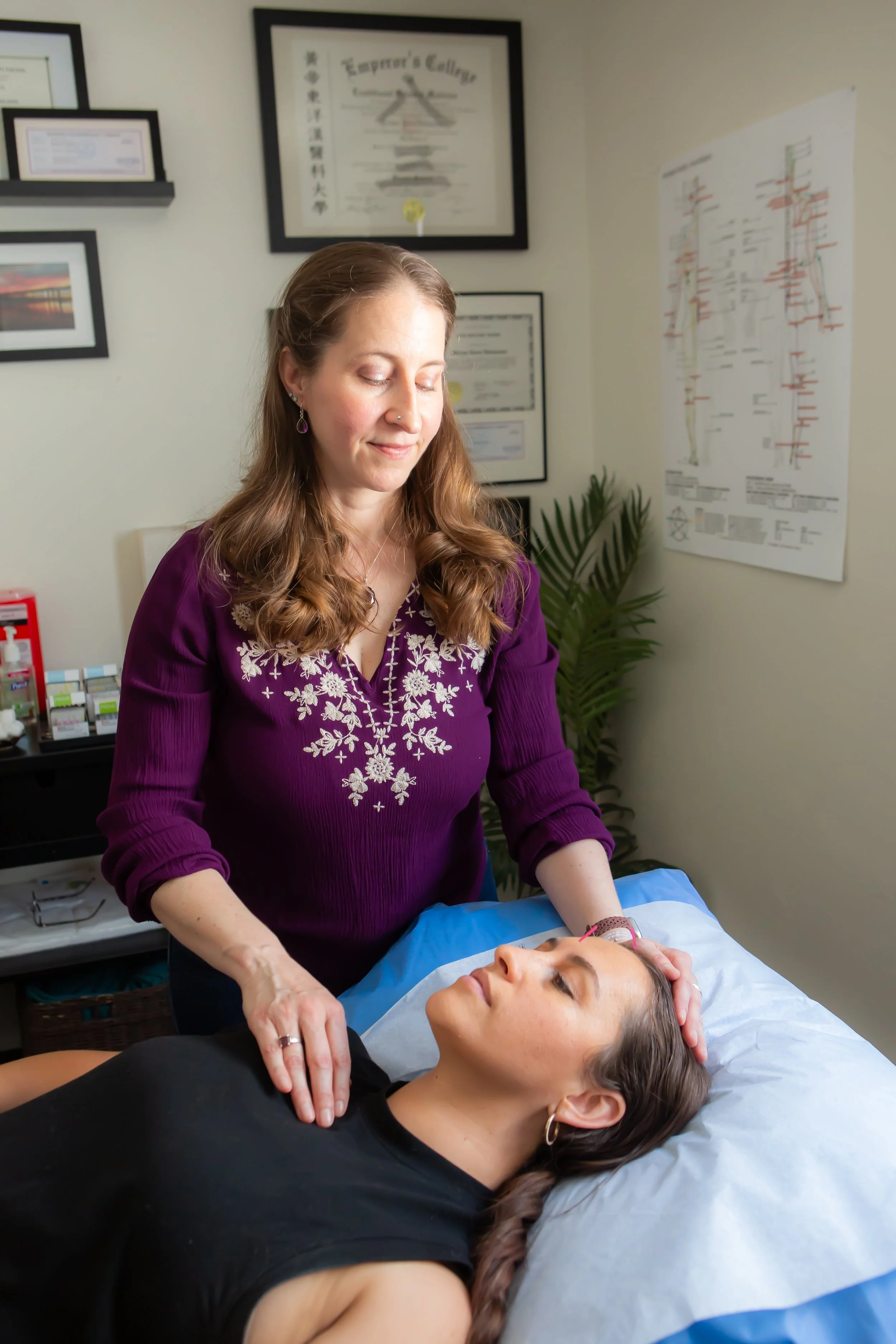A woman in a purple embroidered top performs an acupuncture treatment on a patient lying on a bed with her eyes closed in a treatment room.