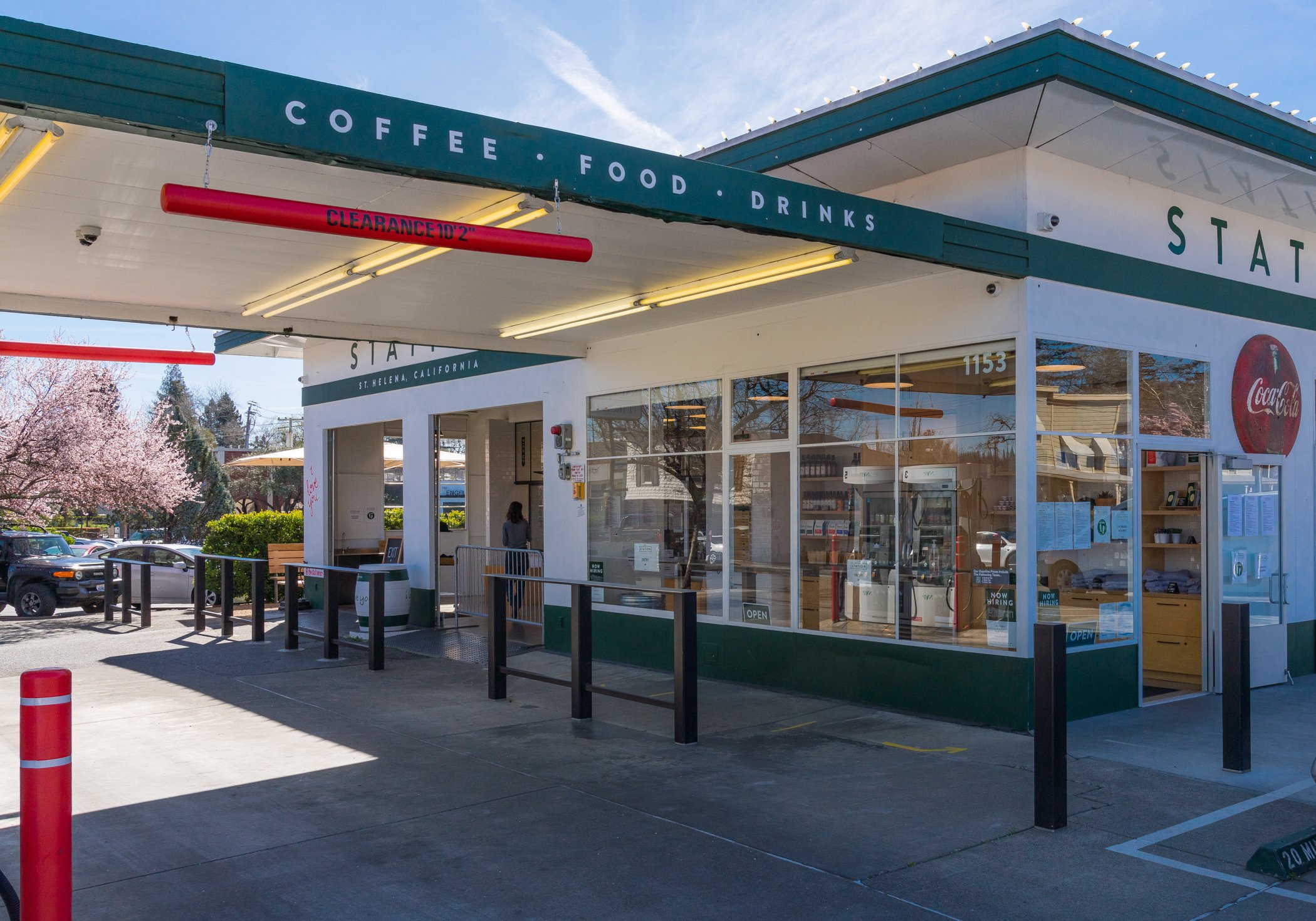 Exterior of a drive-thru coffee shop with a green and white design, displaying signs for coffee, food, and drinks. The store has large glass windows and a Coca-Cola sign. There's a clearance sign in red and a partially visible car parked nearby.