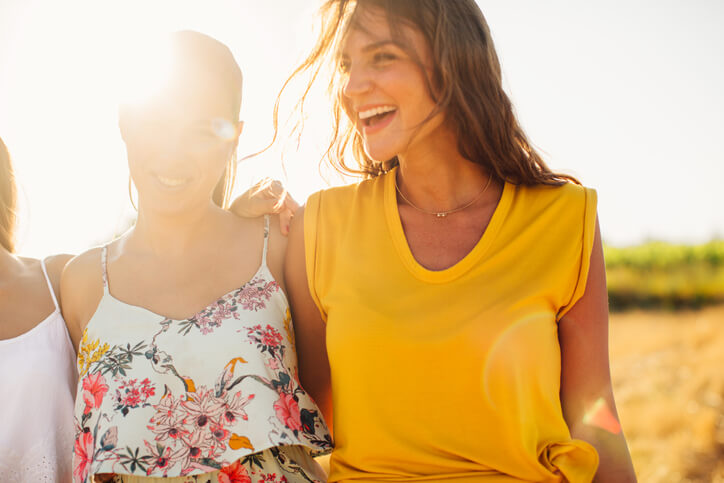 Three women smiling outdoors on a sunny day, with one wearing a yellow sleeveless top and another in a floral dress.