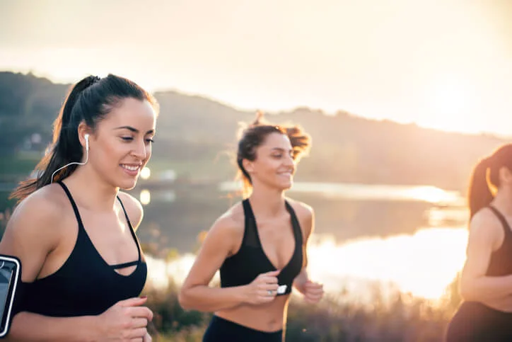 Three women jogging outdoors near a lake during sunset, dressed in black athletic wear, smiling and enjoying the activity.