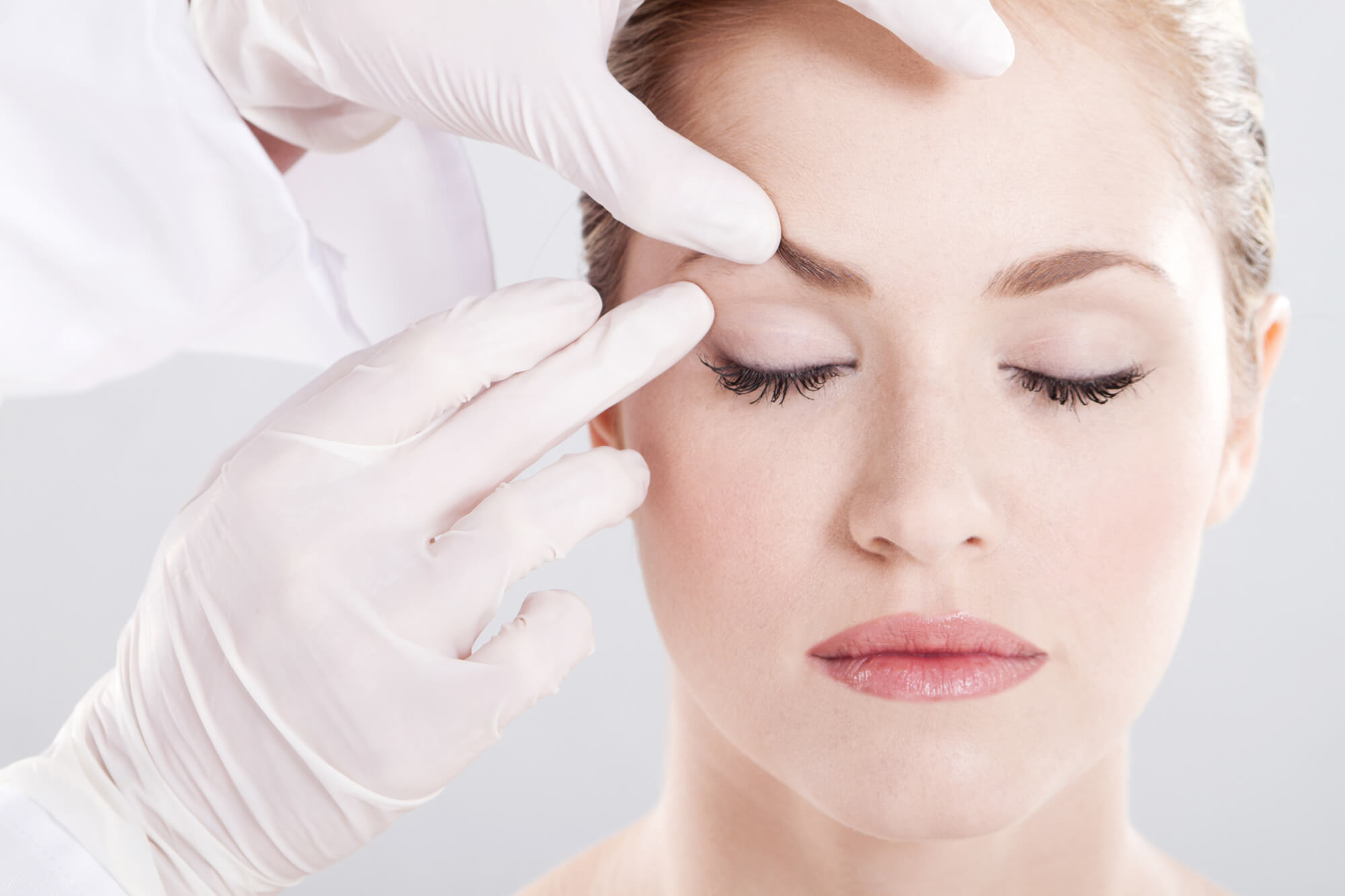 A woman receives a cosmetic treatment around her eyebrows from a professional wearing gloves.
