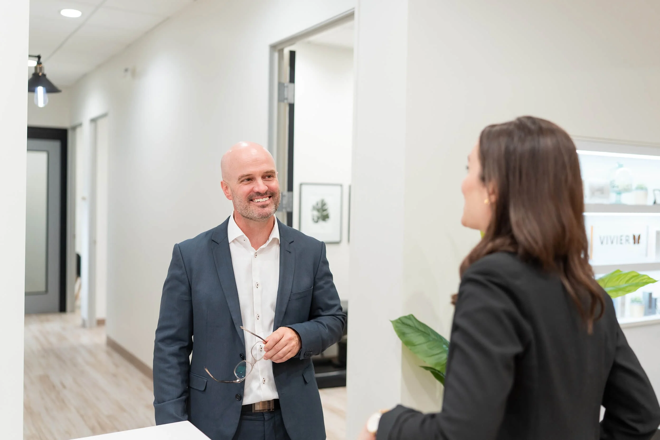 Man smiling and holding glasses while talking to woman in business attire in modern office hallway.