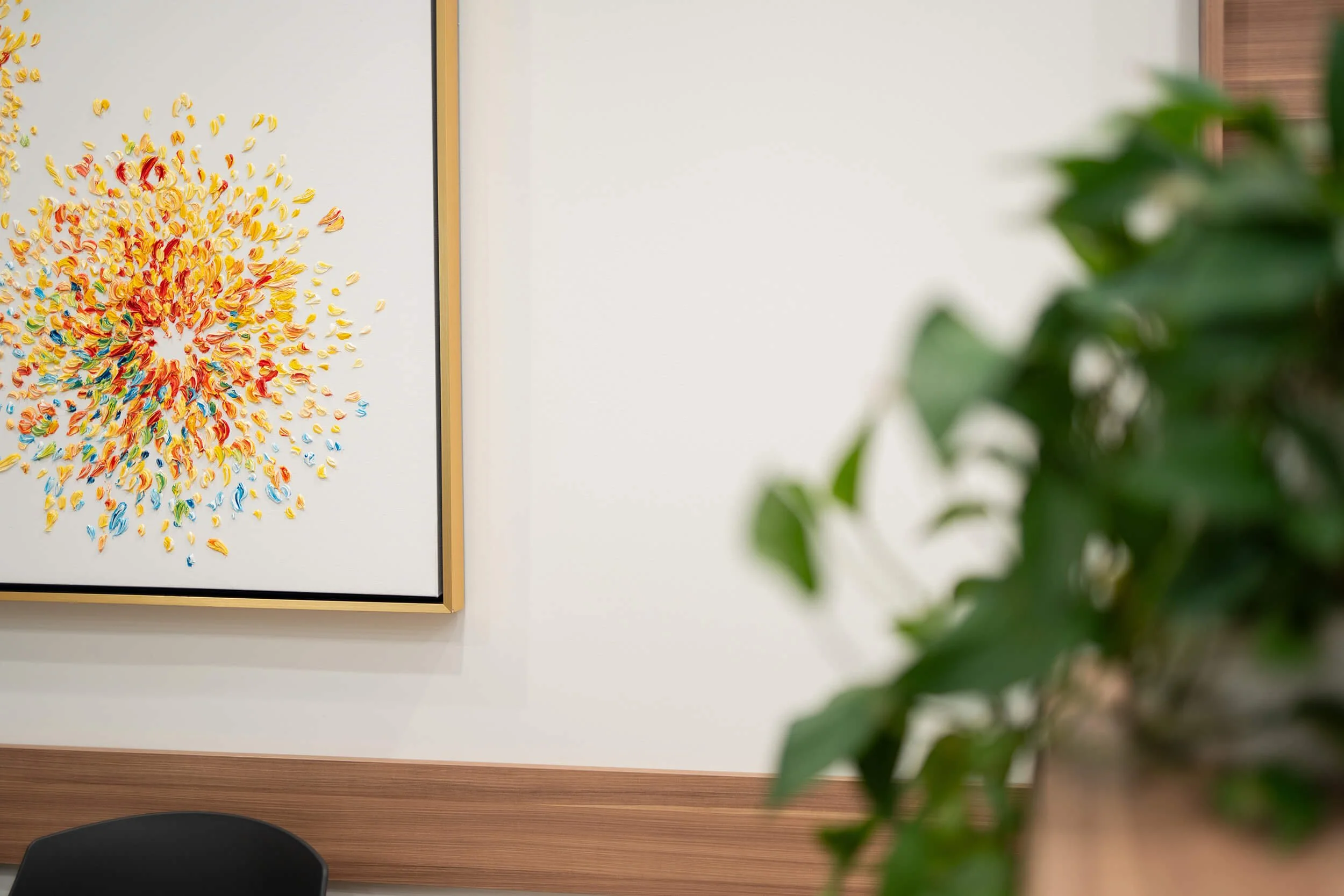 Close-up of a colorful, abstract, textured painting hanging on a white wall with a wooden trim, partially obscured by a green leafy plant in the foreground.