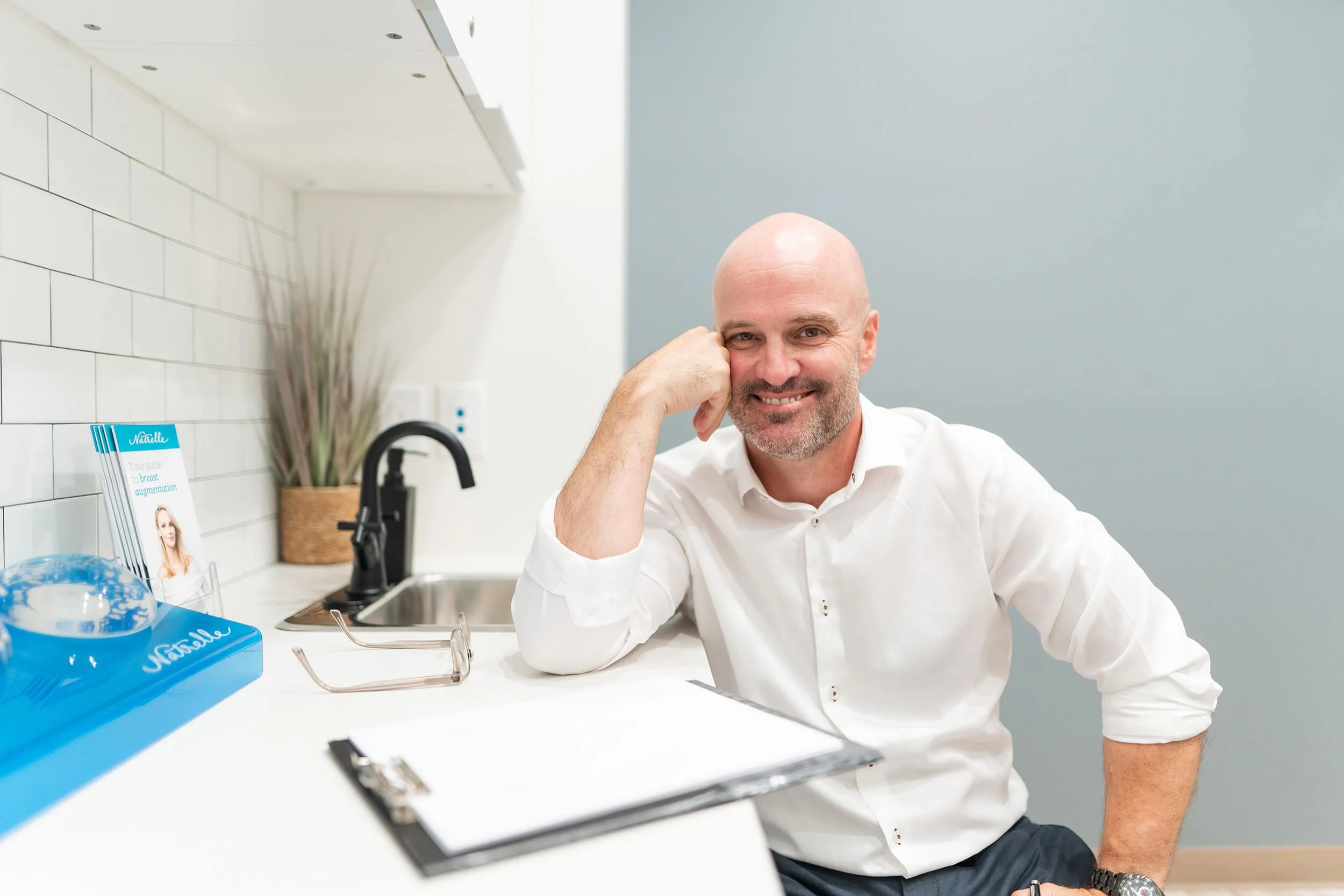 A smiling bald man in a white shirt sitting at a kitchen counter with a clipboard, glasses, and a skincare product display nearby.
