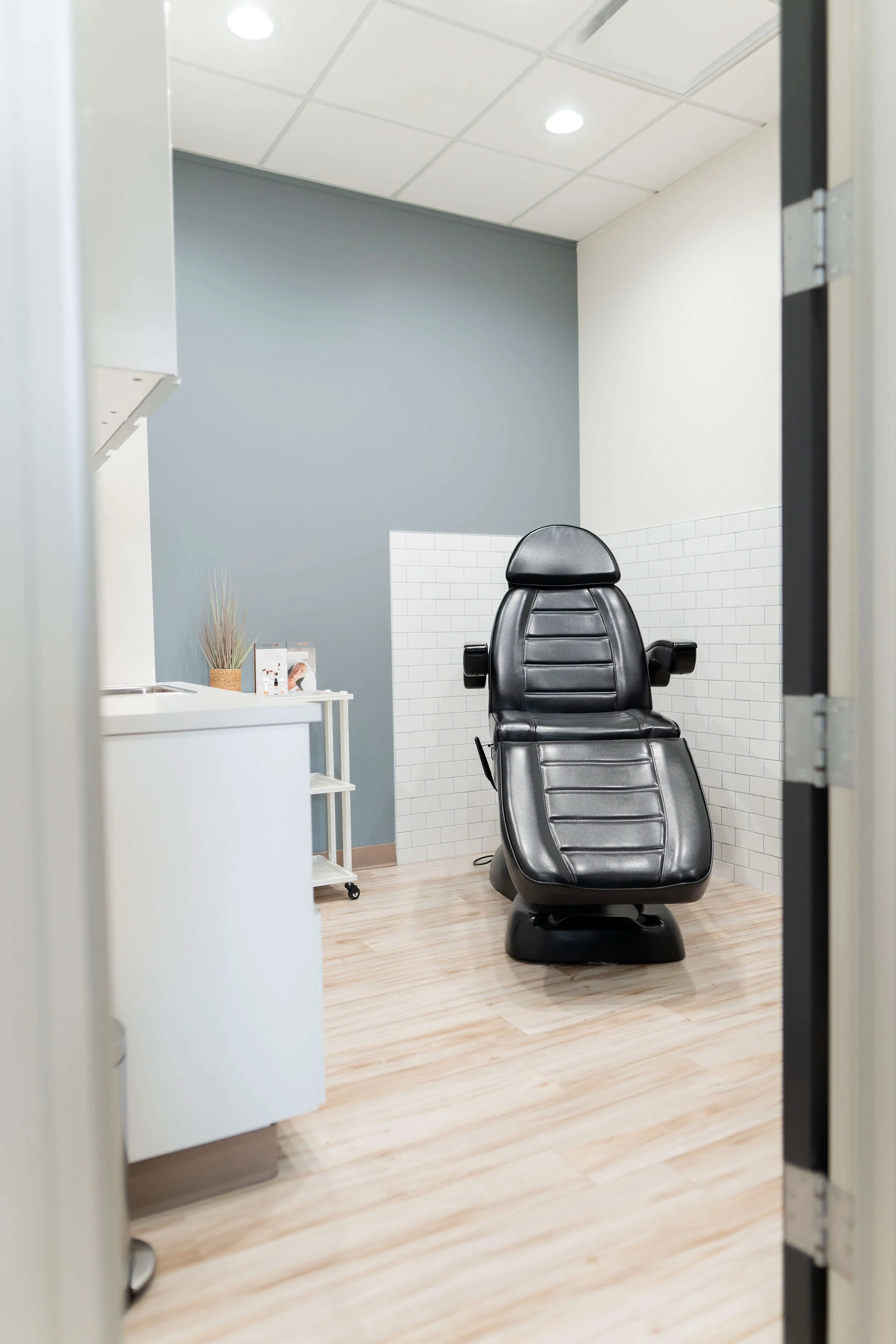 A medical examination room with a black leather chair, white tiled wall, wooden floor, gray and white painted walls, and a small white cart with plants and photo frames.