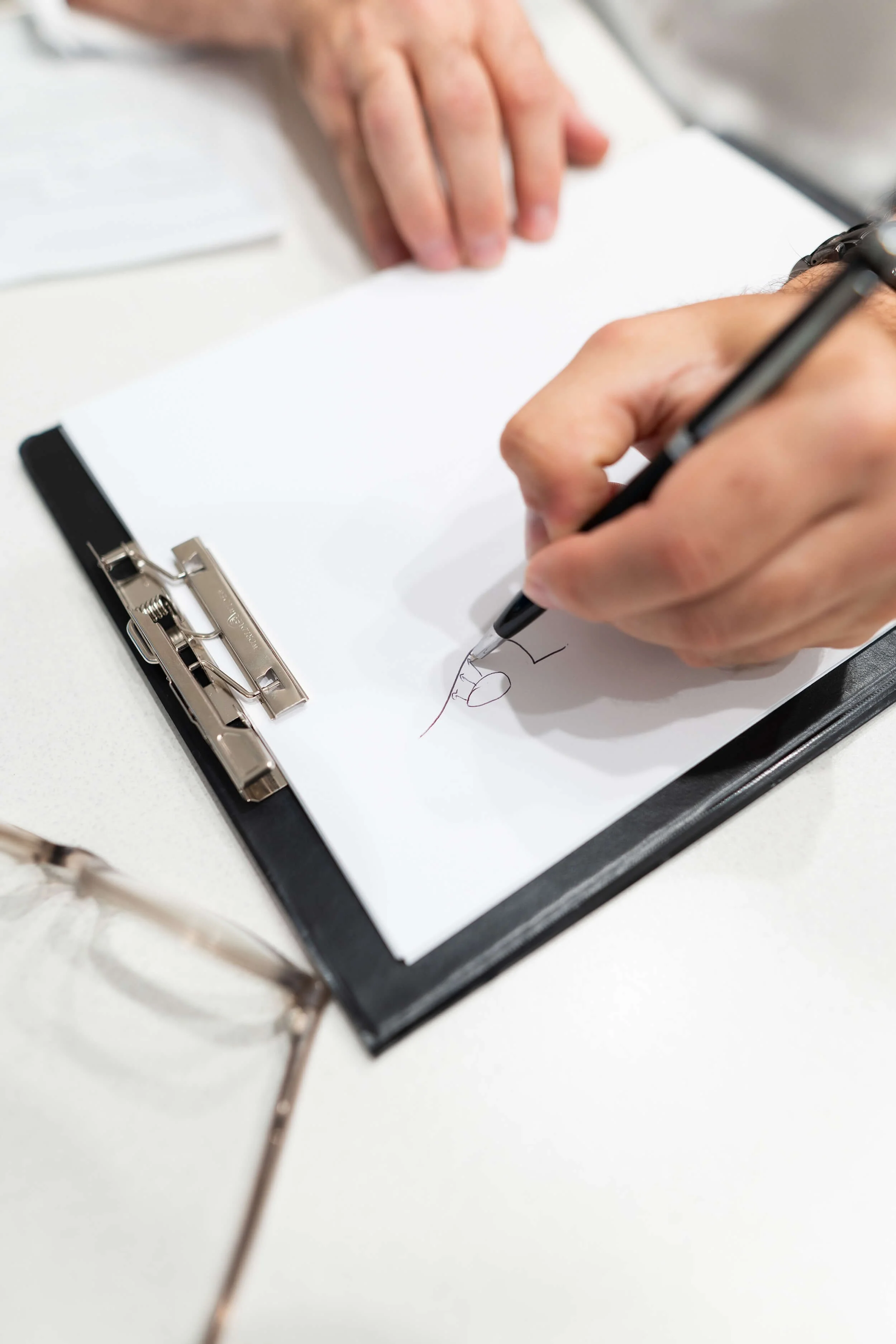 Person writing on a clipboard with a black pen, with their left hand resting on the table, glasses to the side, and some papers in the background.