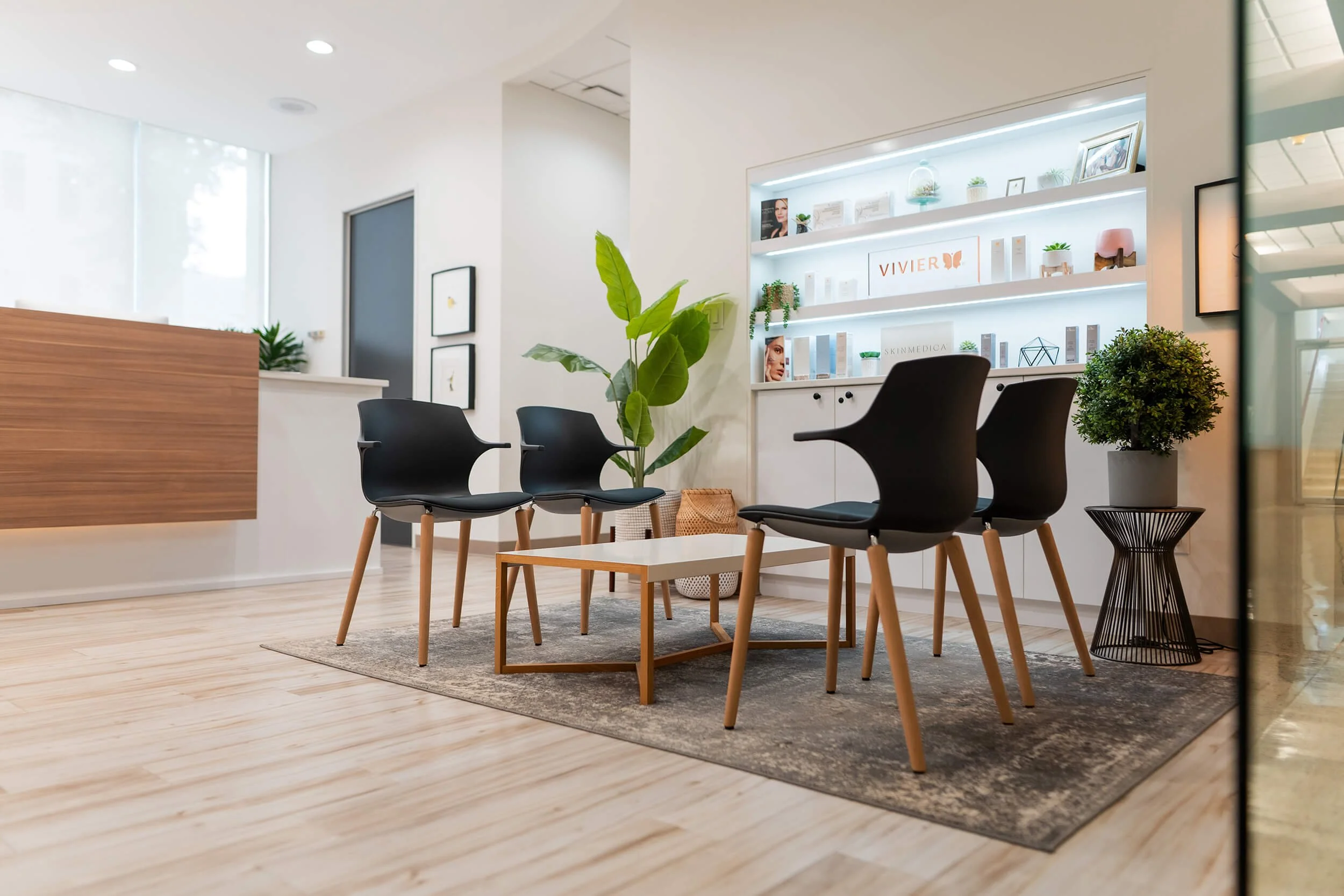 Modern waiting room with black chairs around a white coffee table, potted plants, and a backlit display shelf with skincare products.