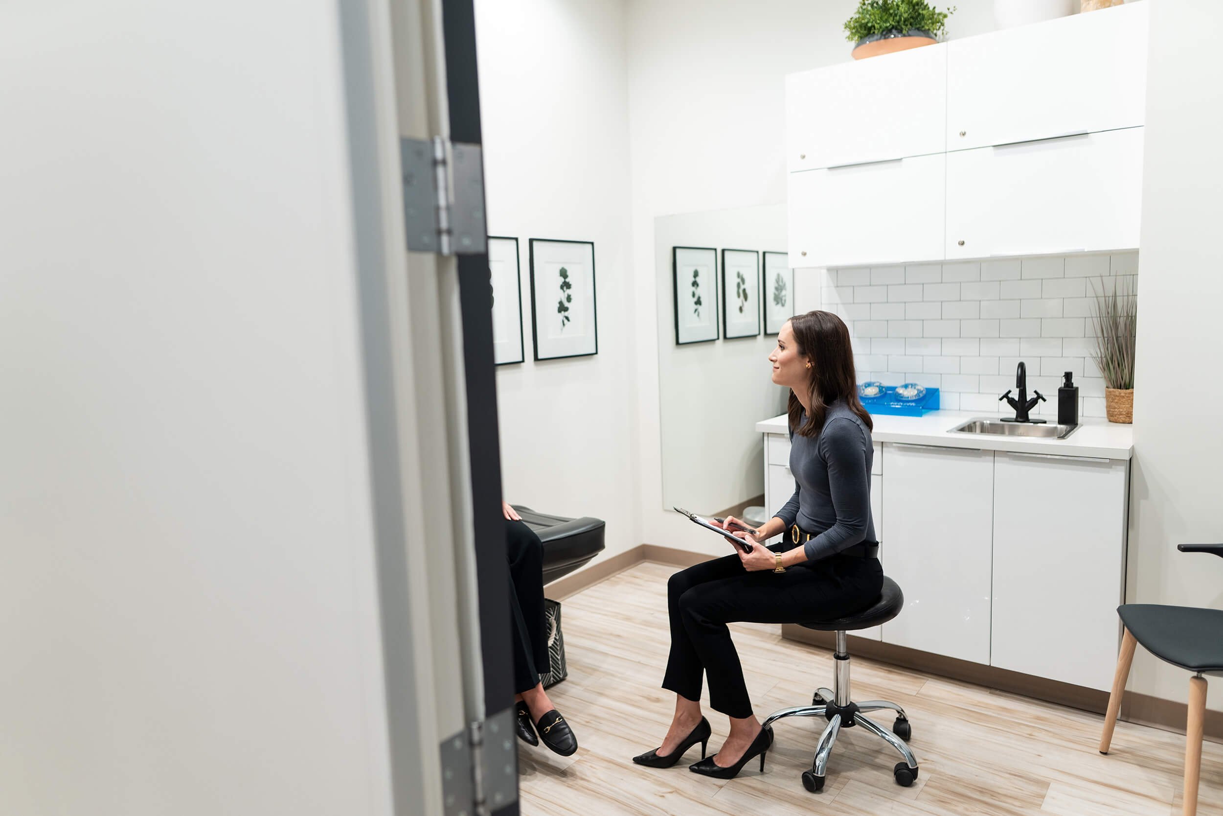A woman sitting on a black stool in a medical office, holding a clipboard and talking to a patient, with framed artwork of plants on the white wall and a white kitchenette in the background.