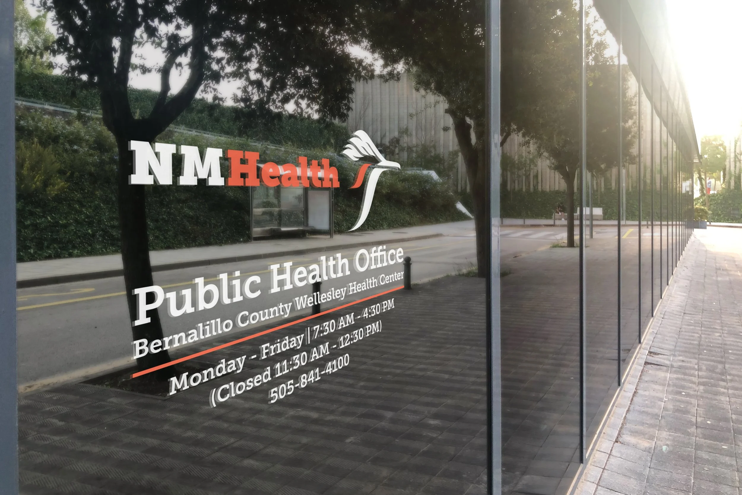 Reflection of trees and a building visible on a glass window with sign for NM Health public health office in Bernalillo County, Wellesley Health Center, with hours of operation listed.