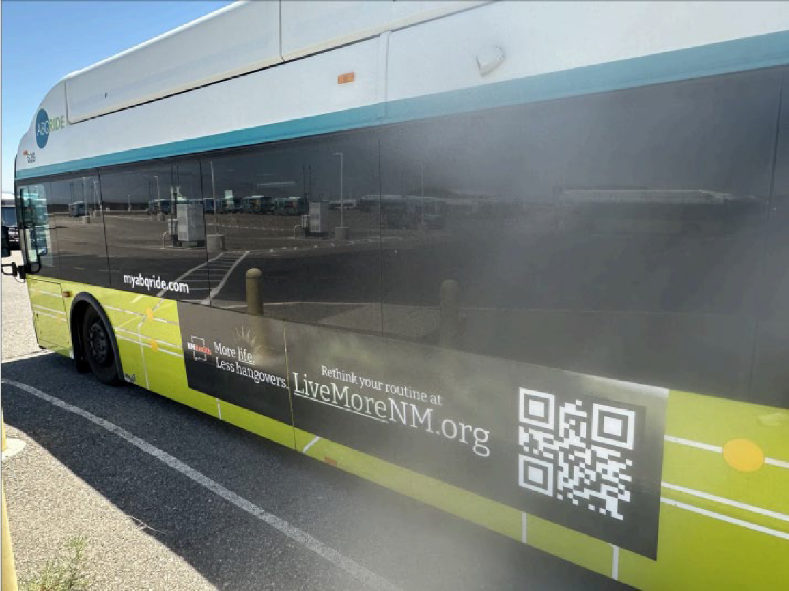 A black bus with green and white graphics parked at a bus stop. The bus displays a QR code and text promoting LiveMoreNM.org, encouraging less hangovers and more life.